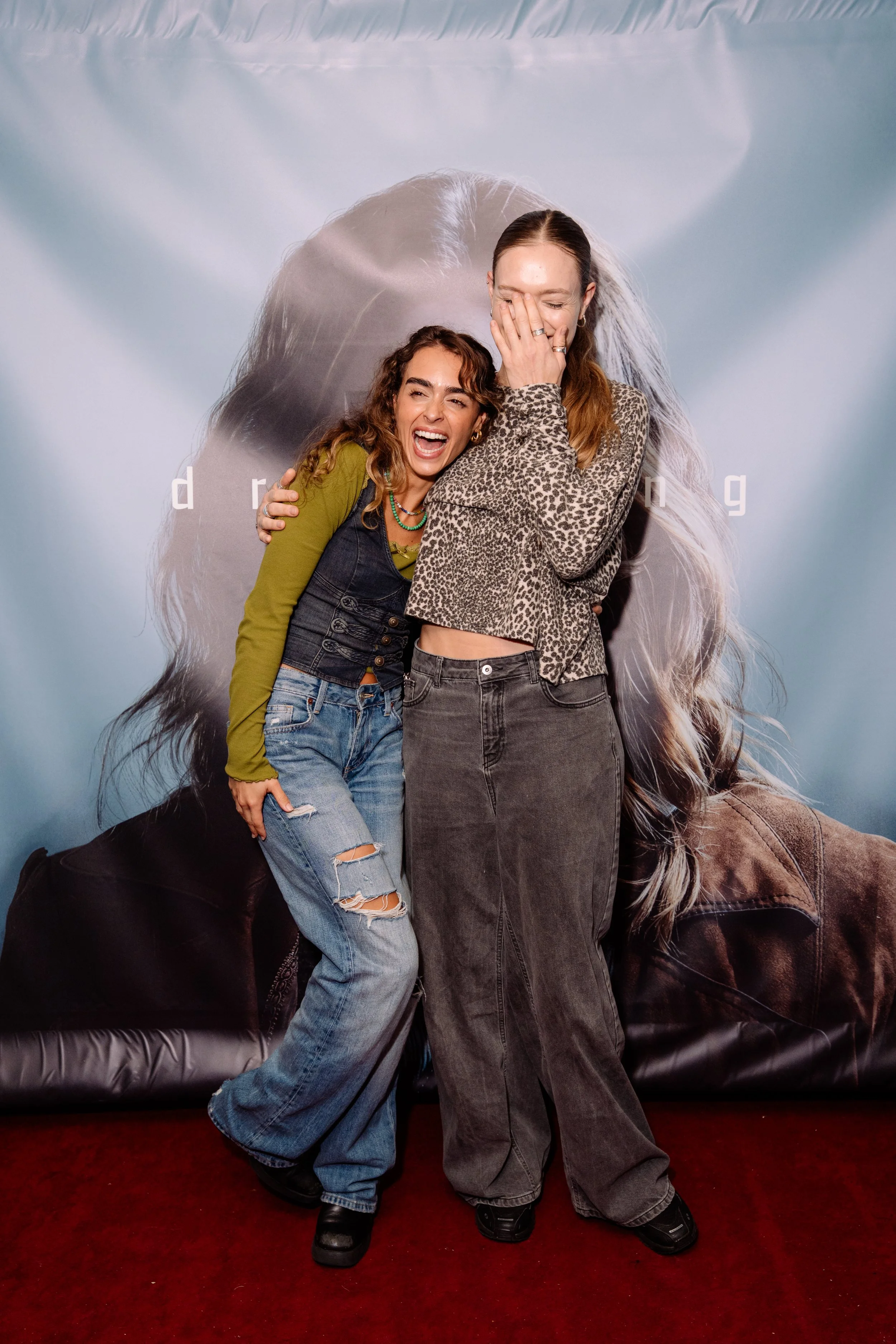 Two women are hugging and smiling in front of a large poster of a woman with long hair. They are standing on a red carpet.