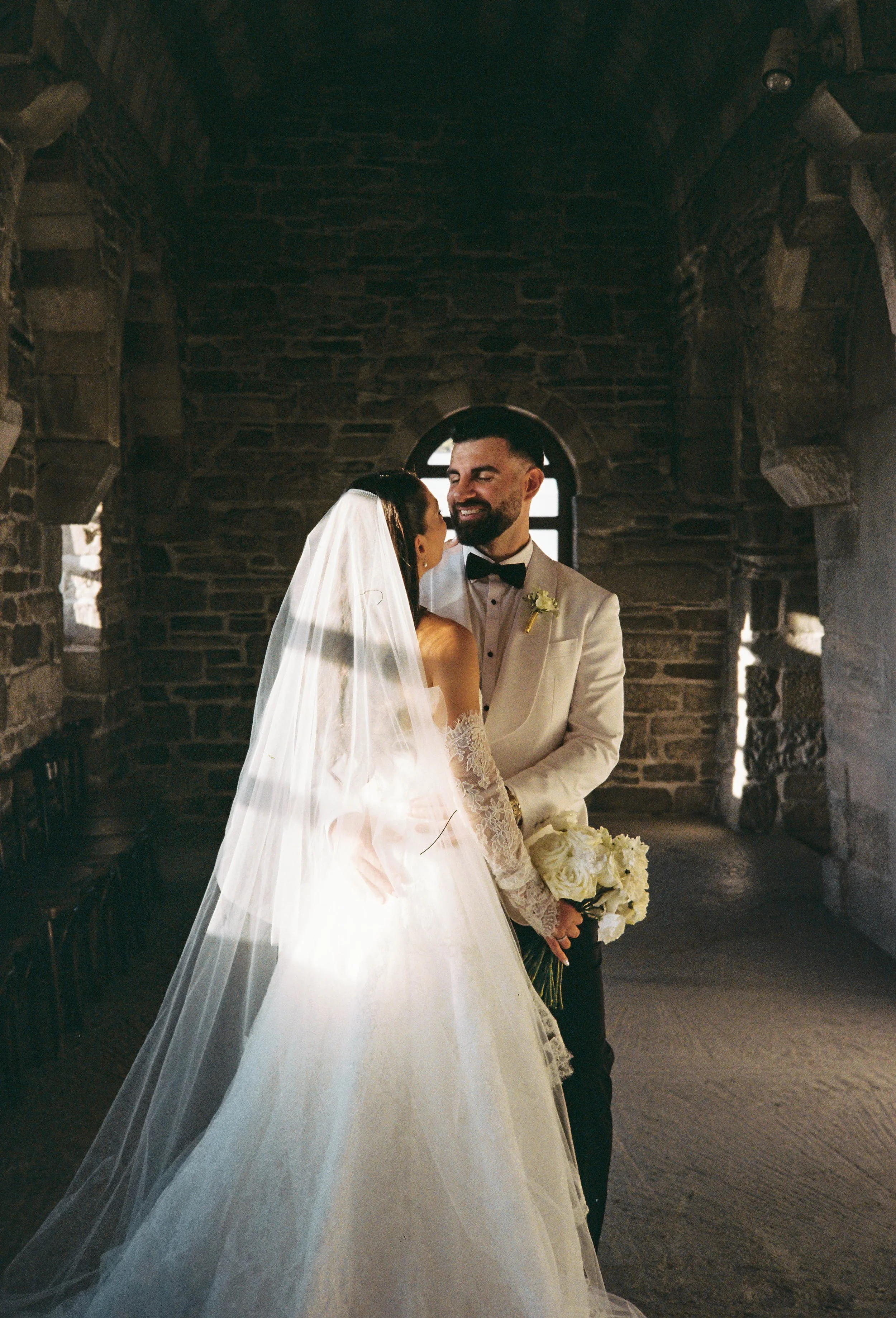 A bride and groom sharing an intimate moment inside a stone-walled building, with the bride holding a bouquet of white flowers.