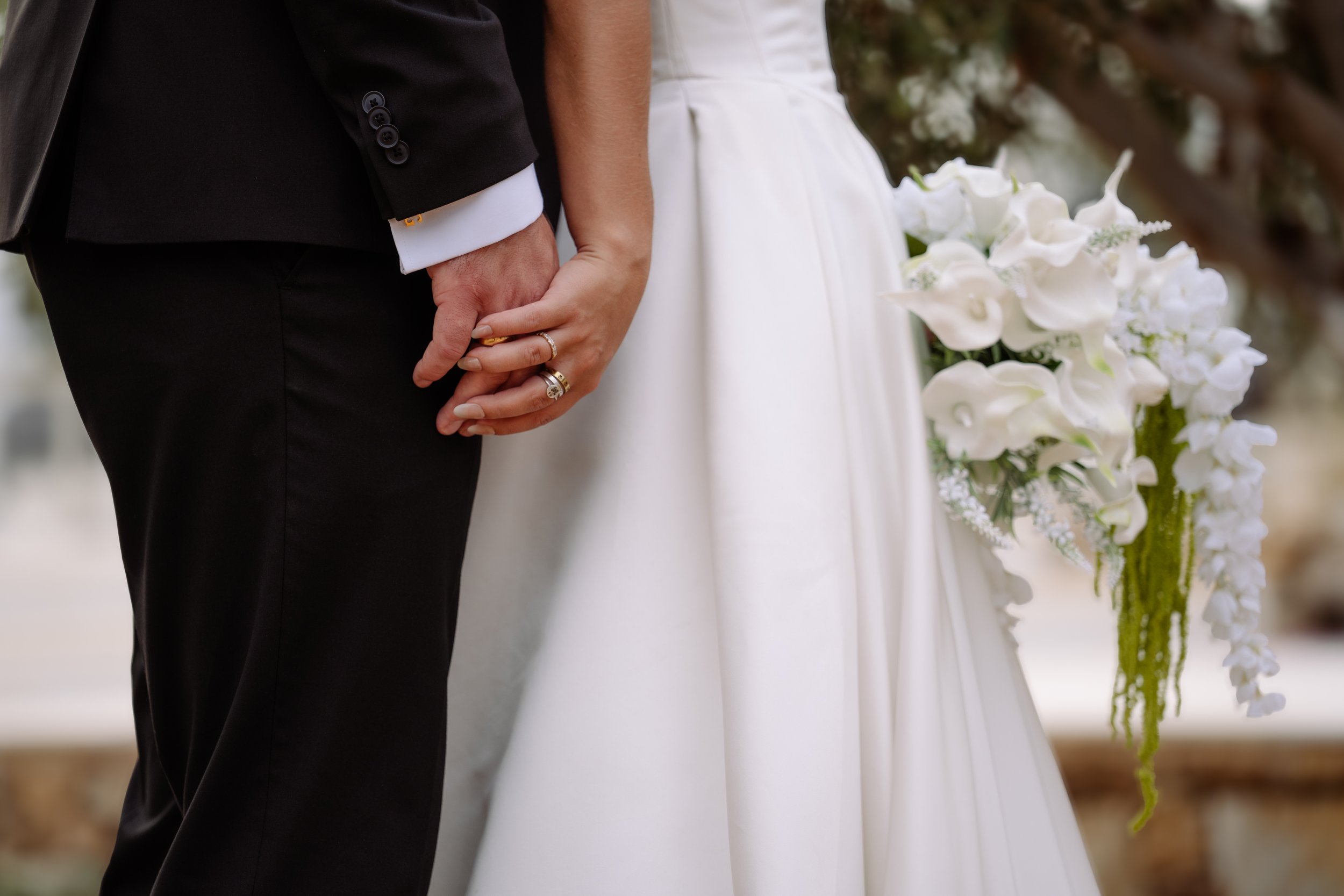 Close-up of a bride and groom holding hands during a wedding ceremony, with the bride wearing a white gown and the groom in a black suit, holding a bouquet of white flowers.