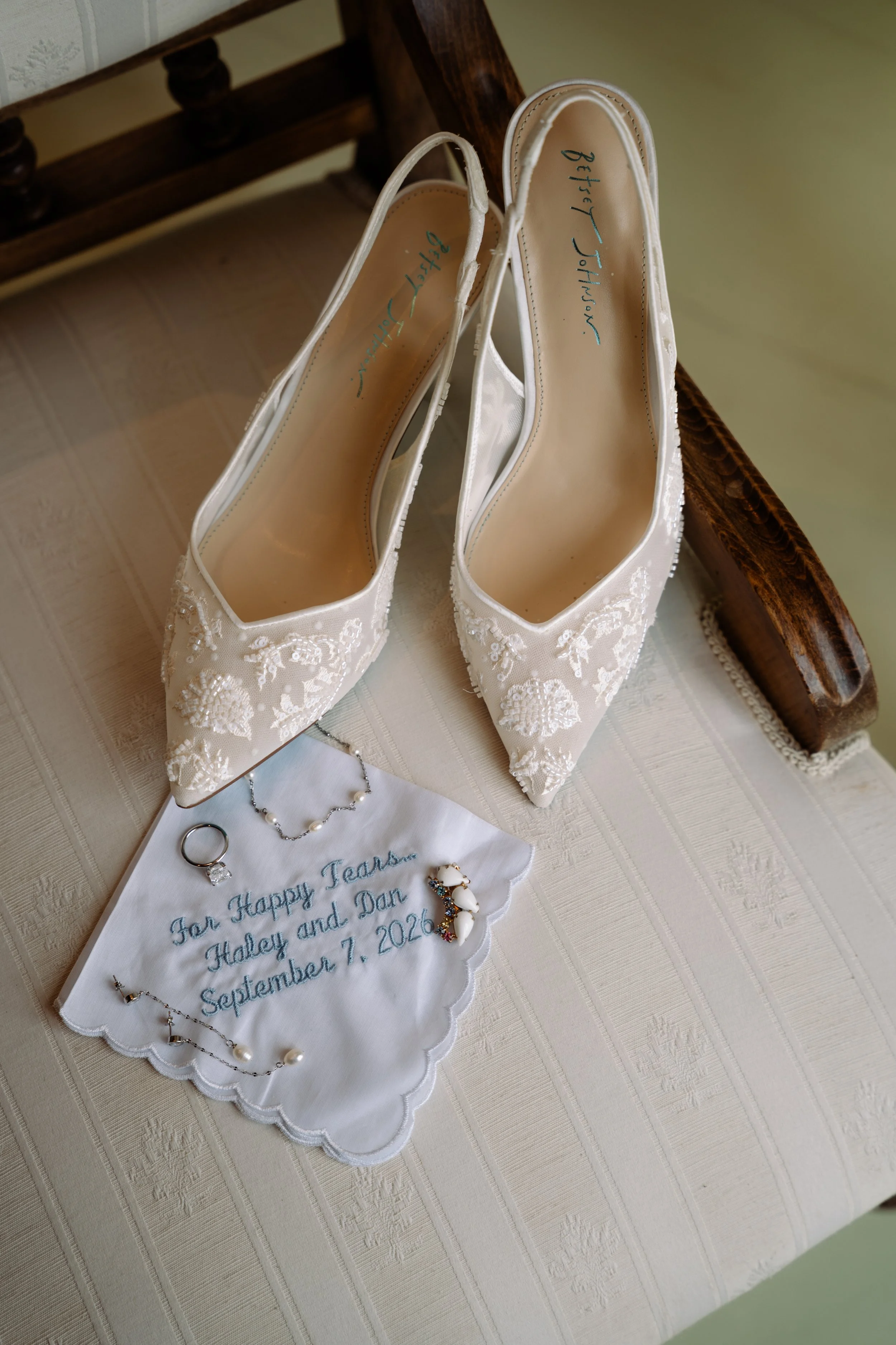 A pair of white bridal shoes with floral embroidery, jewelry including rings, earrings, and a bracelet, along with a cloth that has a wedding message embroidered on it, all placed on a beige tablecloth.