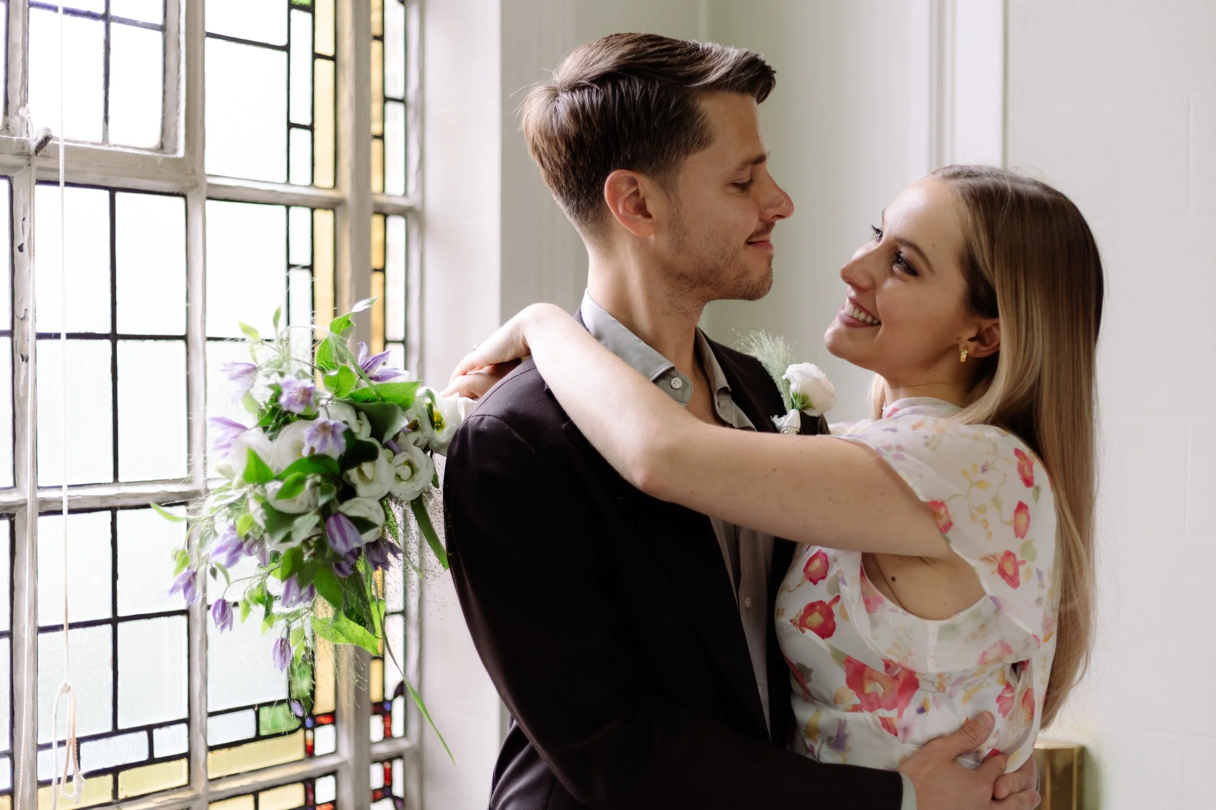 A couple smiling and embracing each other indoors near a stained glass window, with the woman holding a bouquet of flowers.