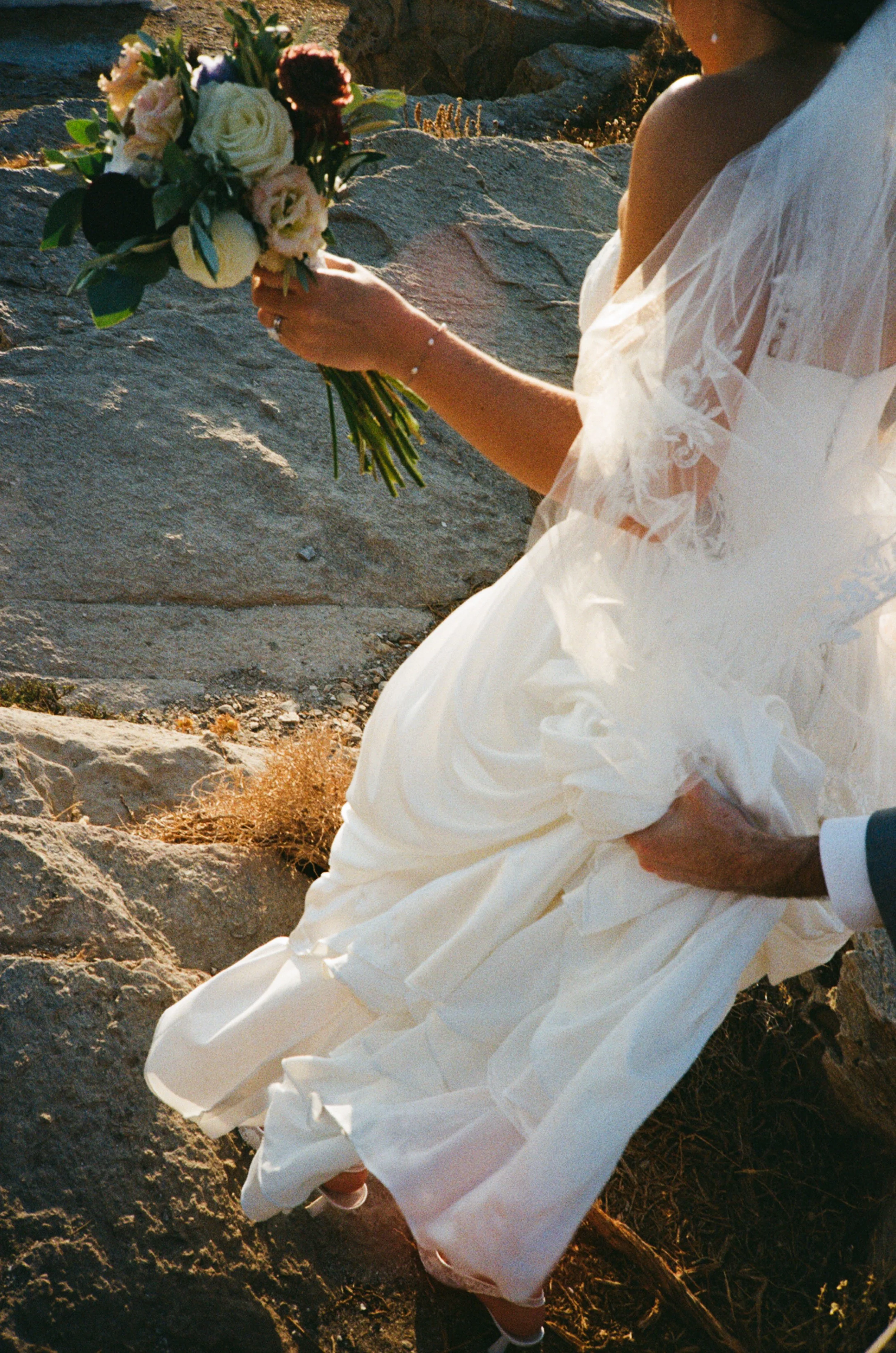 A person in a white wedding dress holding a bouquet of flowers, sitting on rocks at sunset.