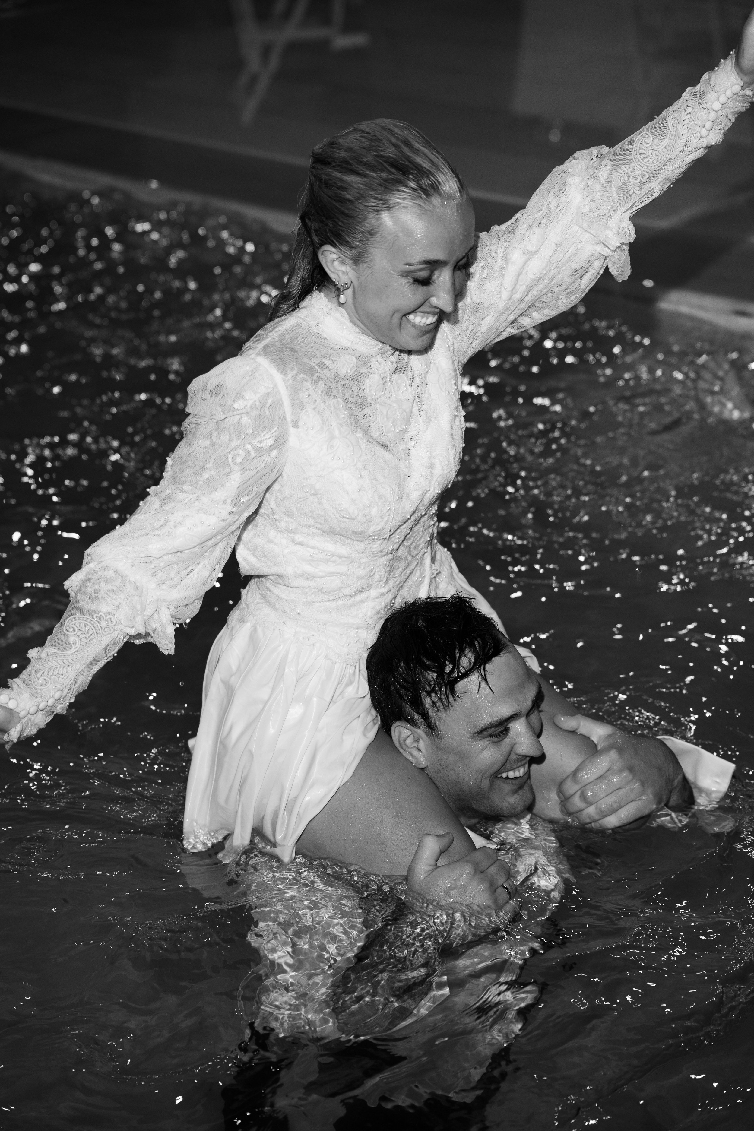 A woman in a lace dress is sitting on a man's shoulders in a pool, smiling and raising her arm, while the man, also smiling, holds her securely in the water.