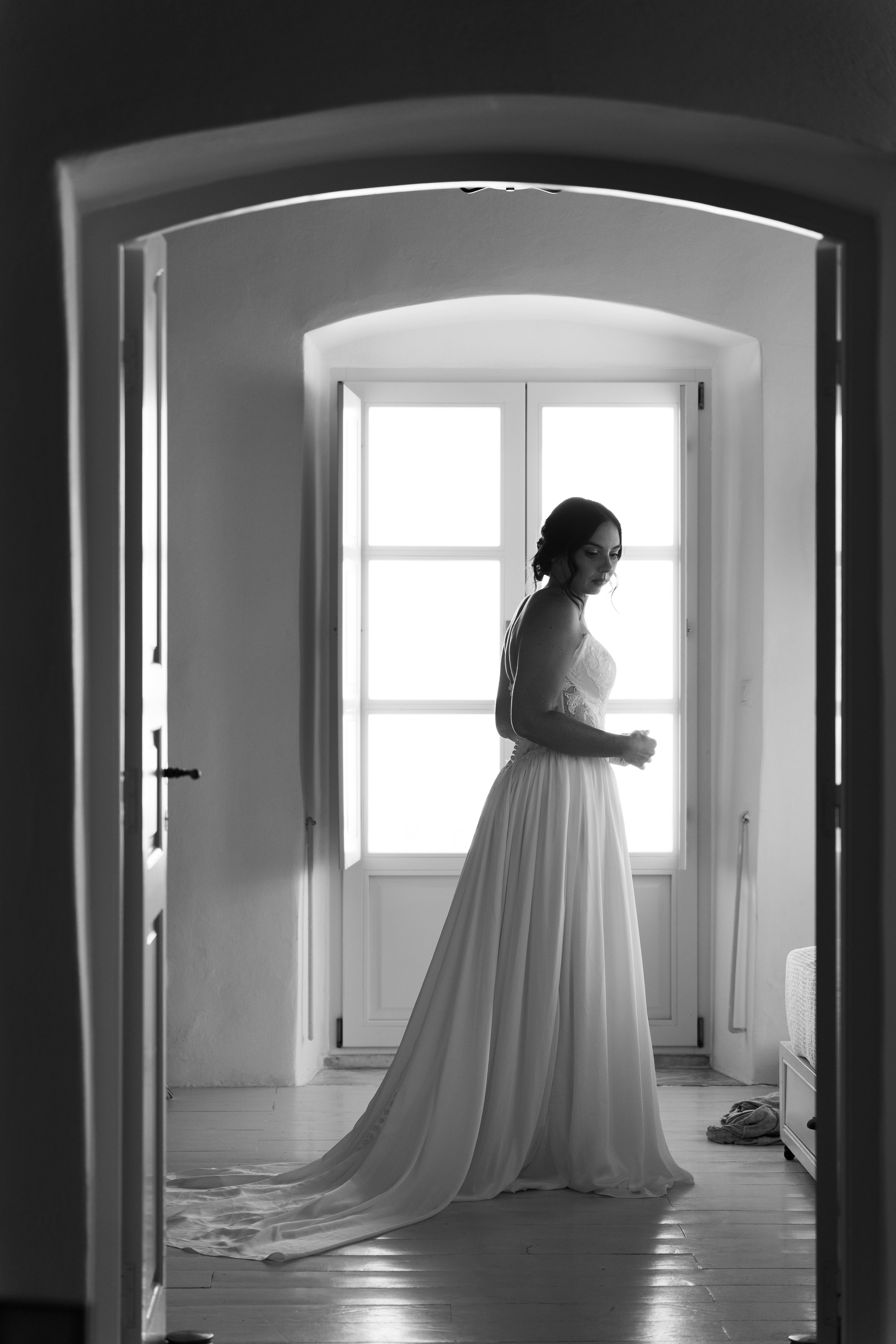 A woman in a wedding dress standing in a sunlit room seen through an arched doorway.