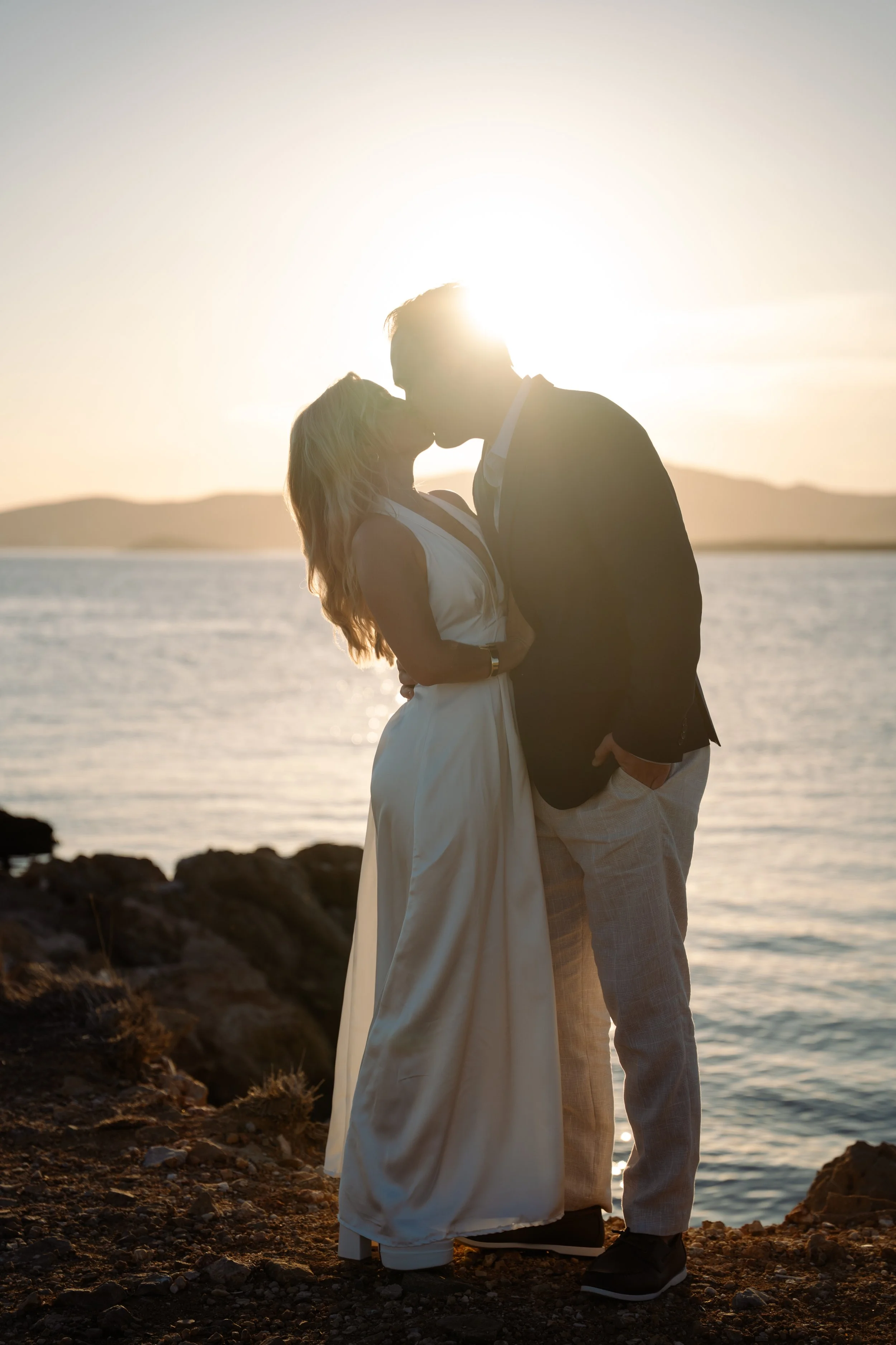 A couple kissing on a rocky shoreline during sunset with a body of water and distant mountains in the background.