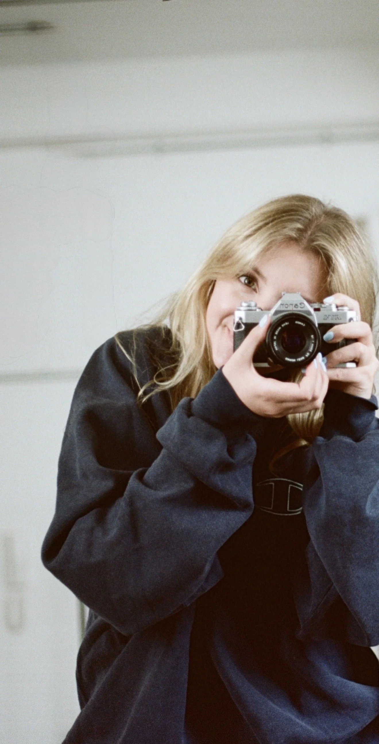A young woman with long blonde hair taking a selfie using a vintage camera in a mirror, wearing a black hoodie.