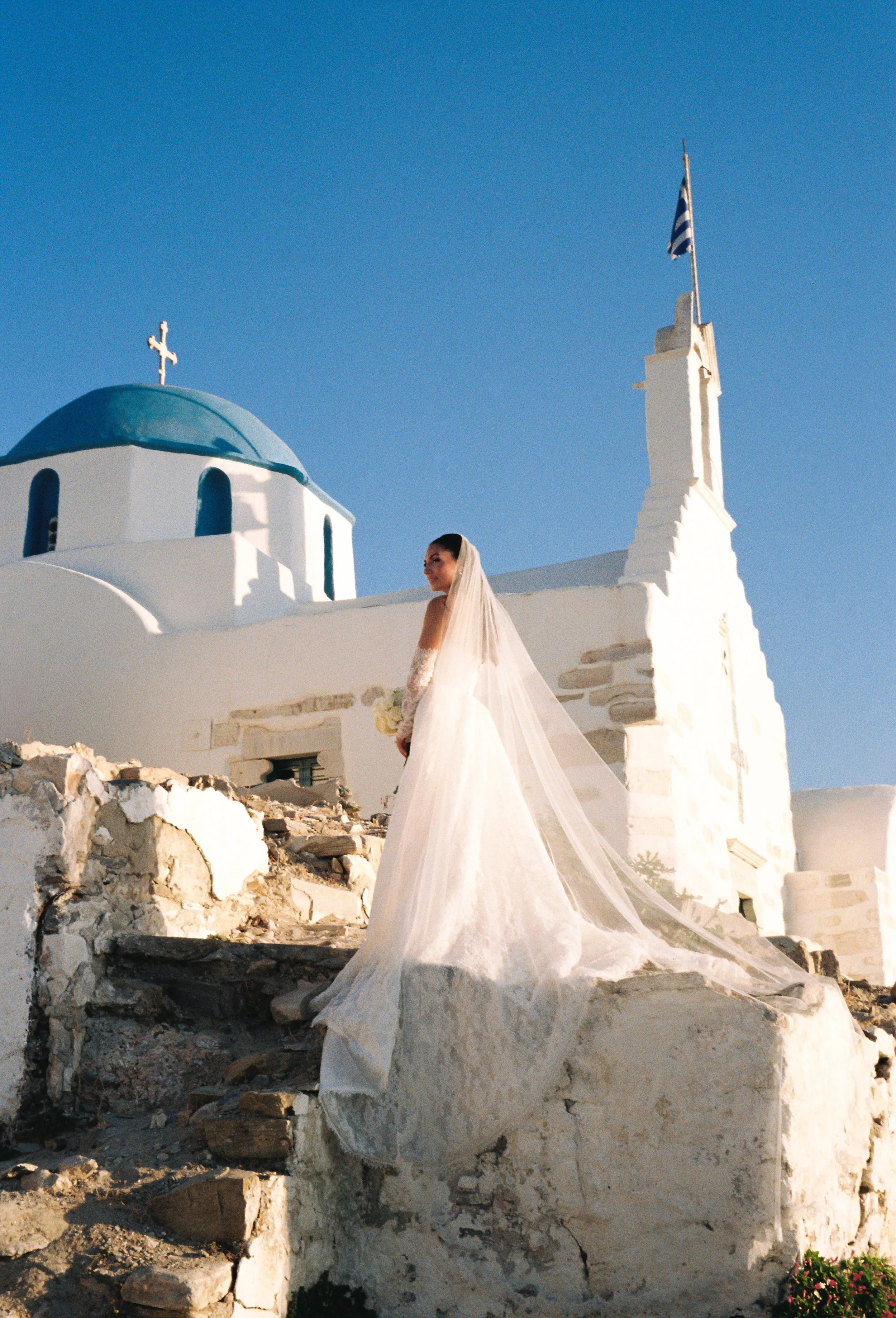 A bride in a wedding dress standing on ancient ruins in front of white buildings with blue domes and a cross, in a Greek island setting.