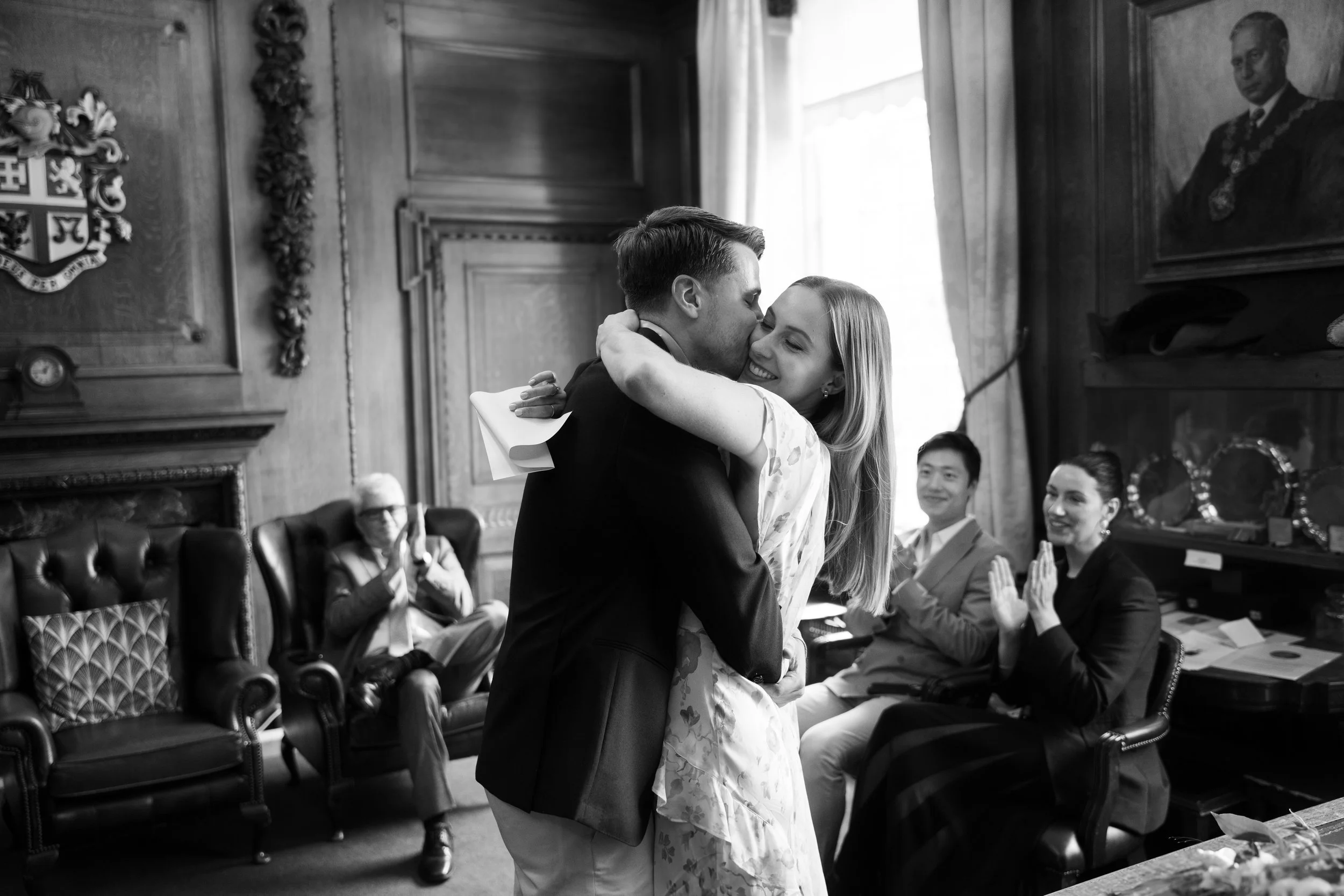 A couple is hugging and kissing during their civil ceremony in central London. Their witnesses are seated and clapping, wearing formal attire.