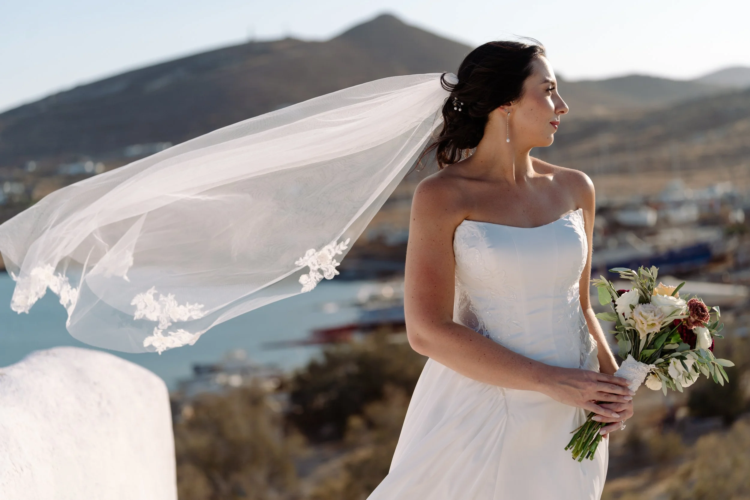 A bride standing outdoors holding a bouquet of flowers, with a flowing veil caught in the wind, overlooking a scenic landscape with hills and water.