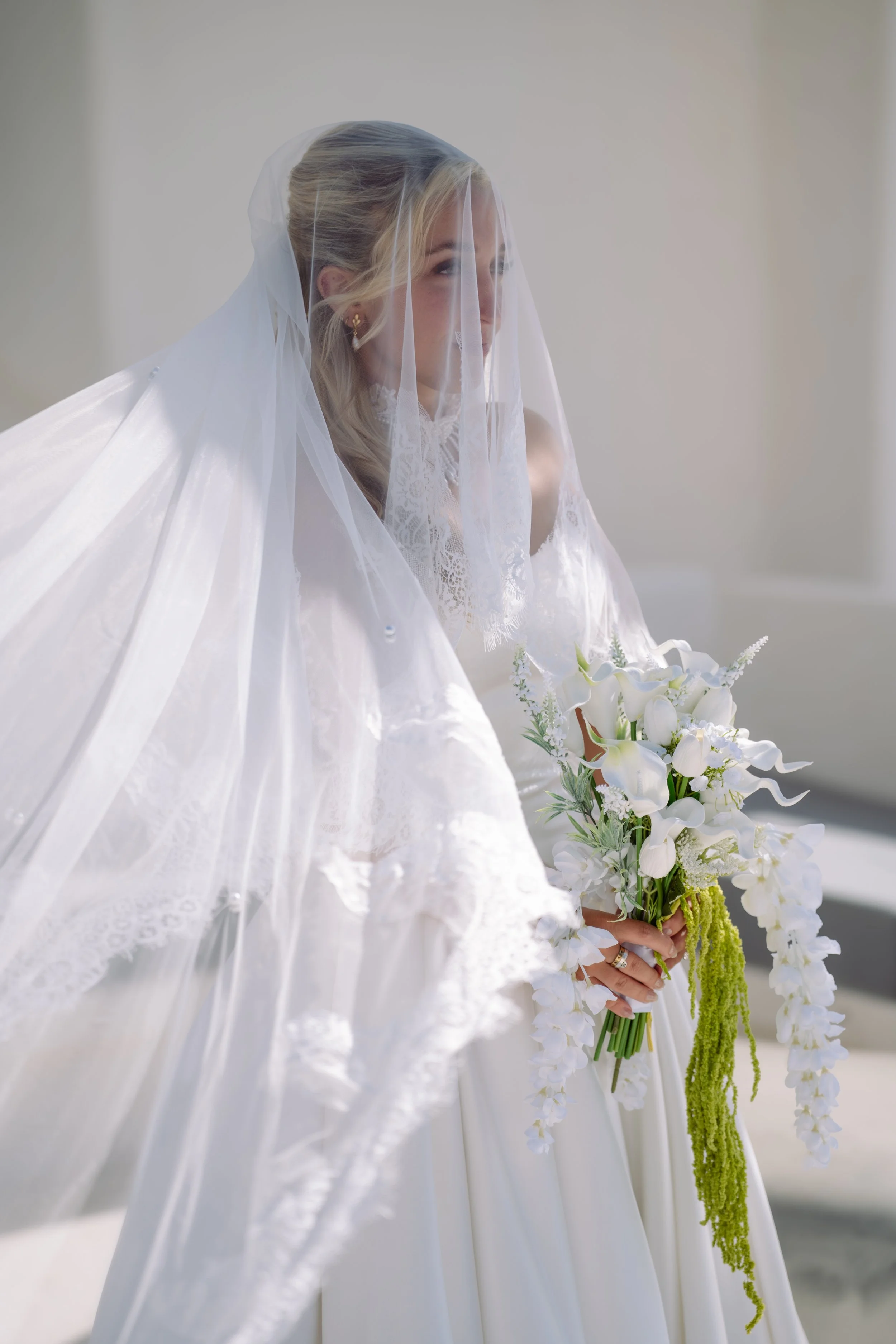 A bride dressed in a white wedding gown with lace details, wearing a sheer veil over her blonde hair styled in loose waves, holding a bouquet of white flowers and greenery, standing indoors against a plain light-colored wall.