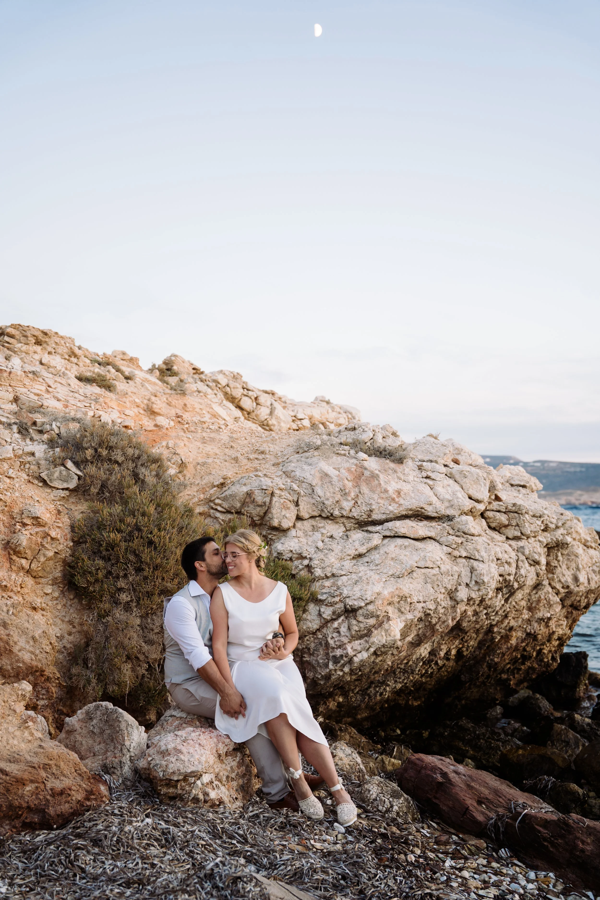 A couple sitting on rocks at a rocky shore, with the man kissing the woman on the cheek, both dressed in light-colored clothing, against a background of large rocks, a bright blue sky, and a visible moon.