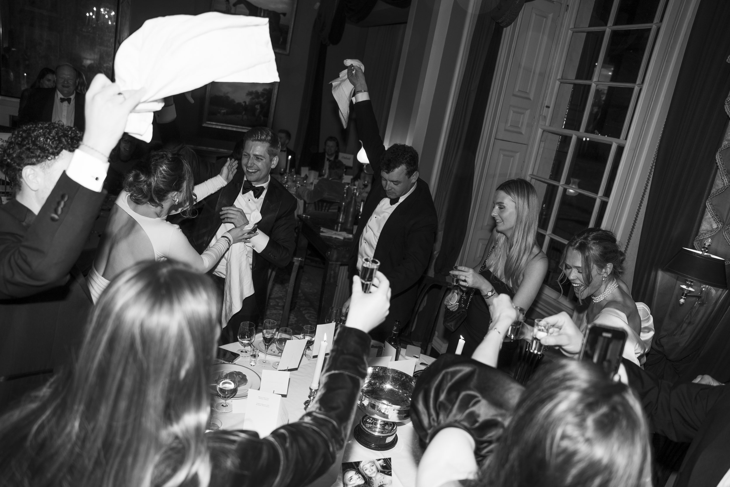 People celebrate at a formal dinner event, wearing tuxedos and elegant dresses, with some raising glasses and others toasting or smiling.