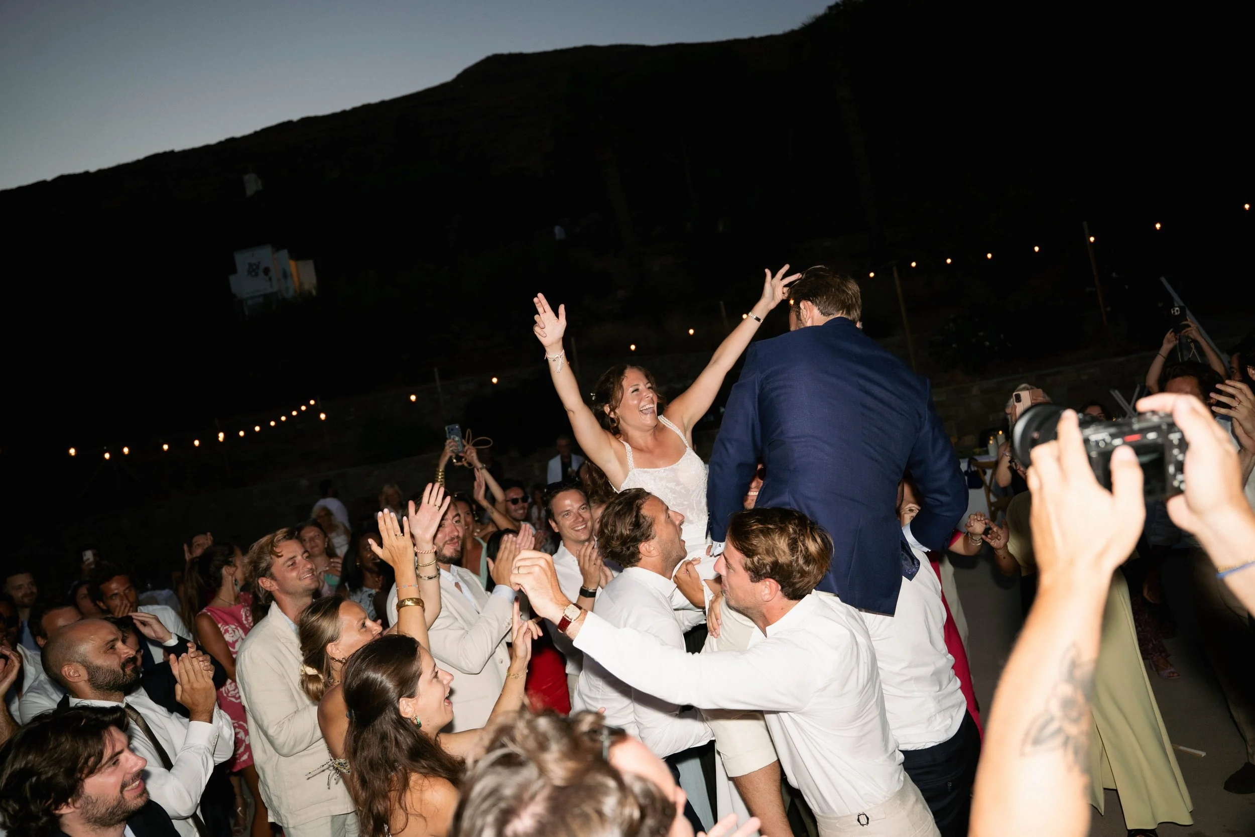 A bride and groom celebrate with guests at their wedding reception, with the bride smiling joyfully and raising her hands in celebration while crowd members cheer around them.