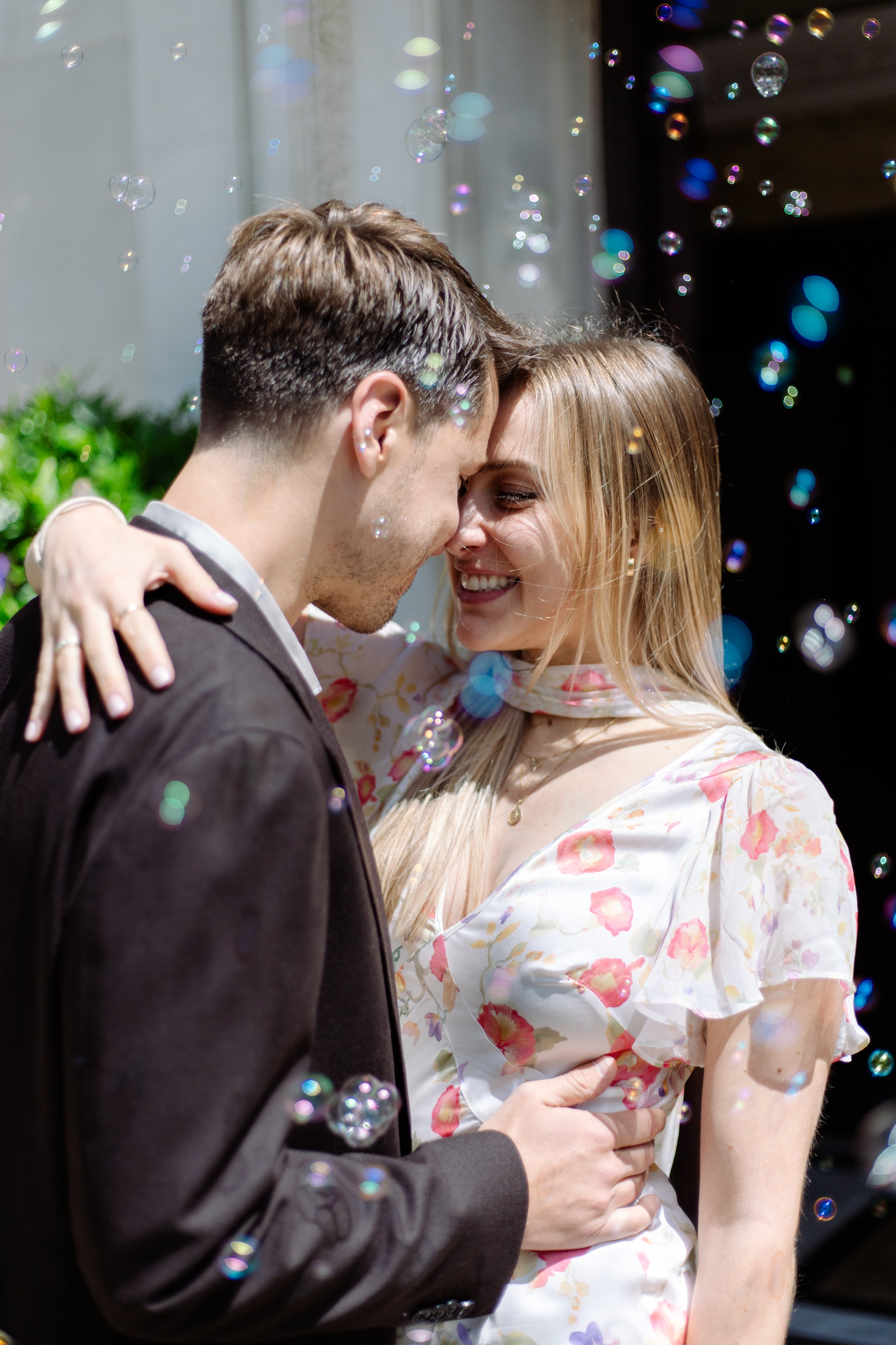 A couple hugging, with the man in a dark suit and the woman in a floral dress, surrounded by floating soap bubbles.