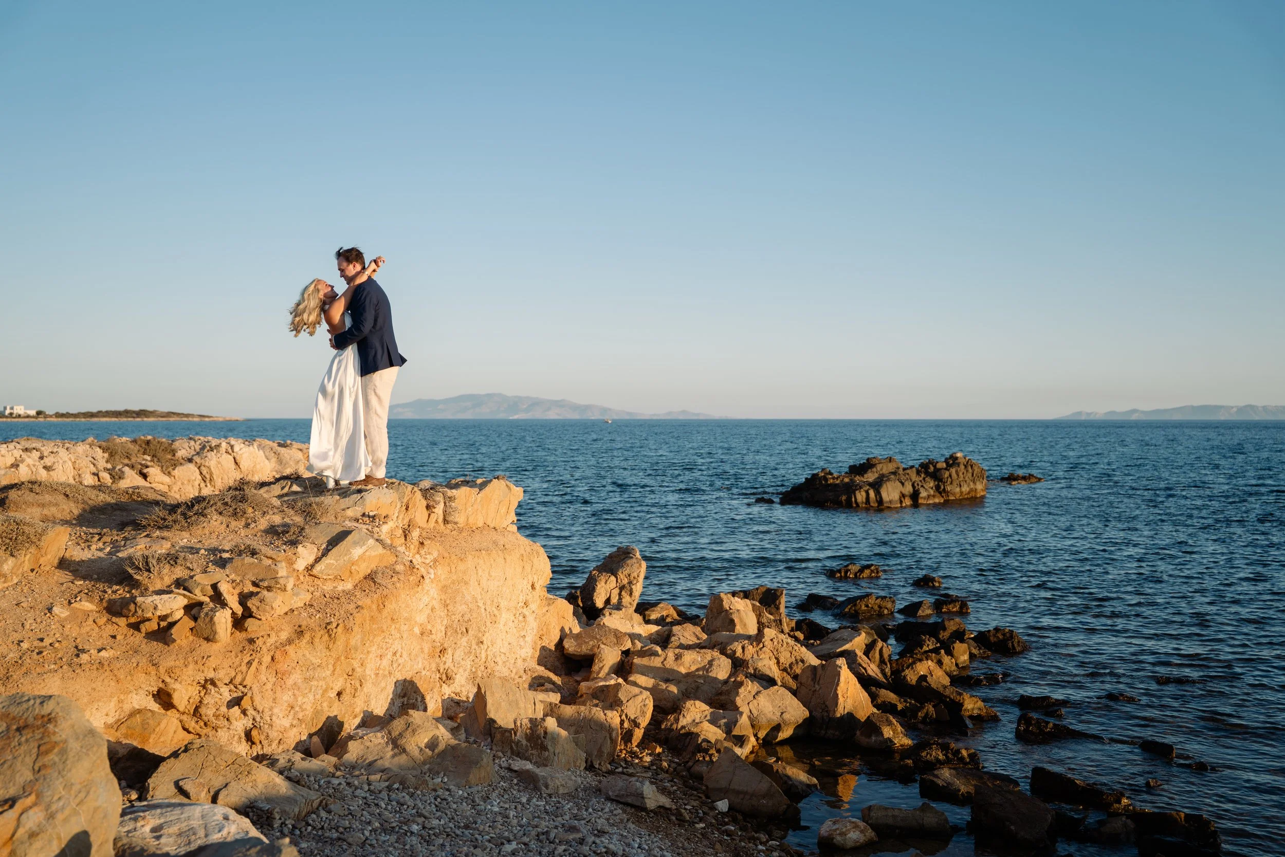 A couple dressed in formal attire standing on rocky shoreline by the ocean, with mountains in the background and clear blue sky.