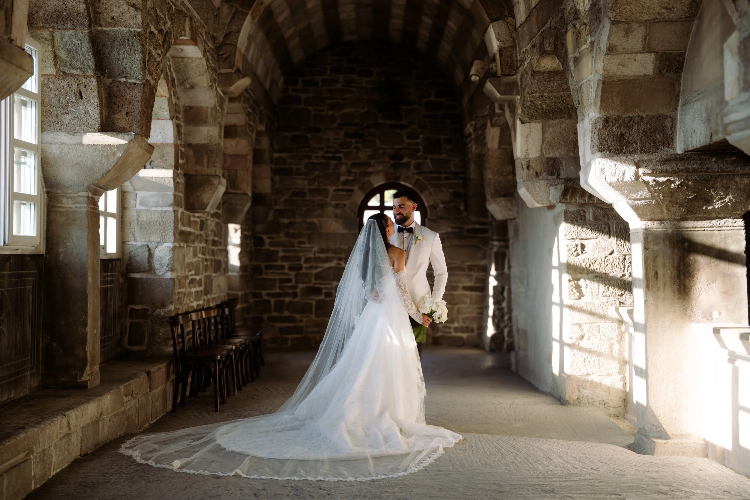 A bride and groom standing in a stone archway, smiling and gazing at each other, during their wedding celebration.