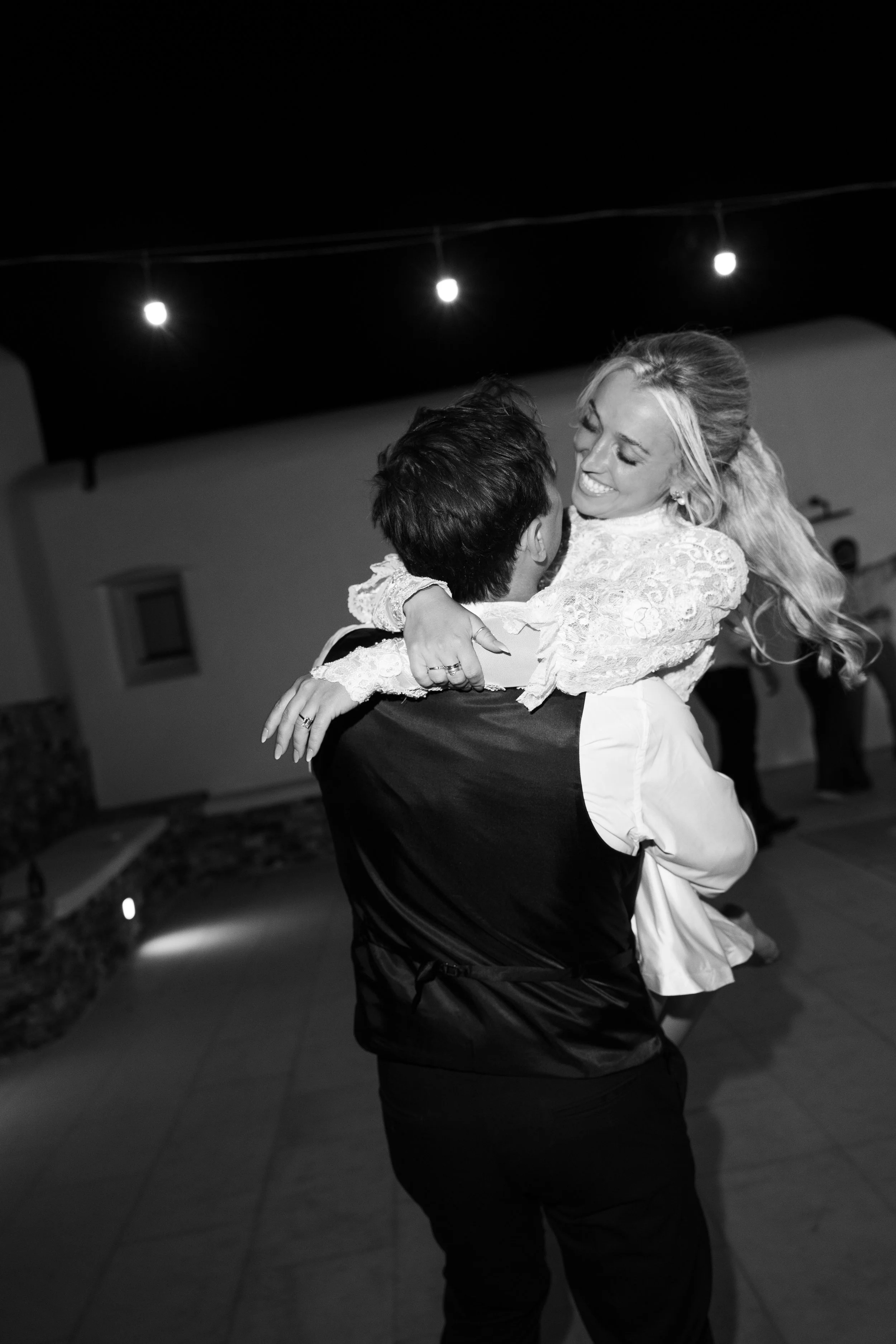 A couple dancing at night outdoors. The woman is smiling and wearing a white lace dress, and the man is holding her while dancing. String lights are visible in the background.