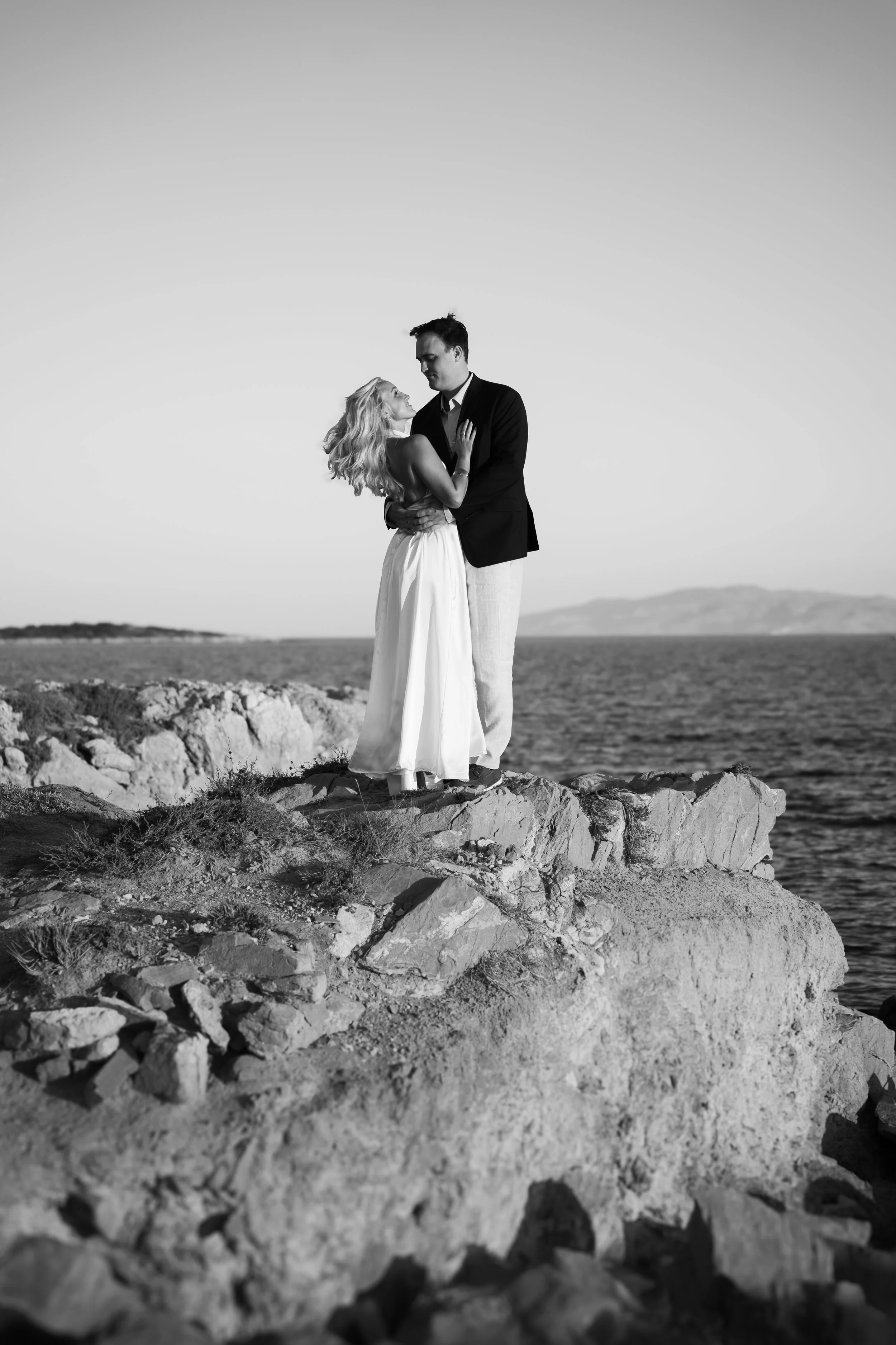 A black and white photo of a couple standing on rocky ground near the ocean, with hills in the background. The woman has long, wavy hair and is wearing a sleeveless dress, while the man is dressed in a blazer and light-colored pants. They are embraci