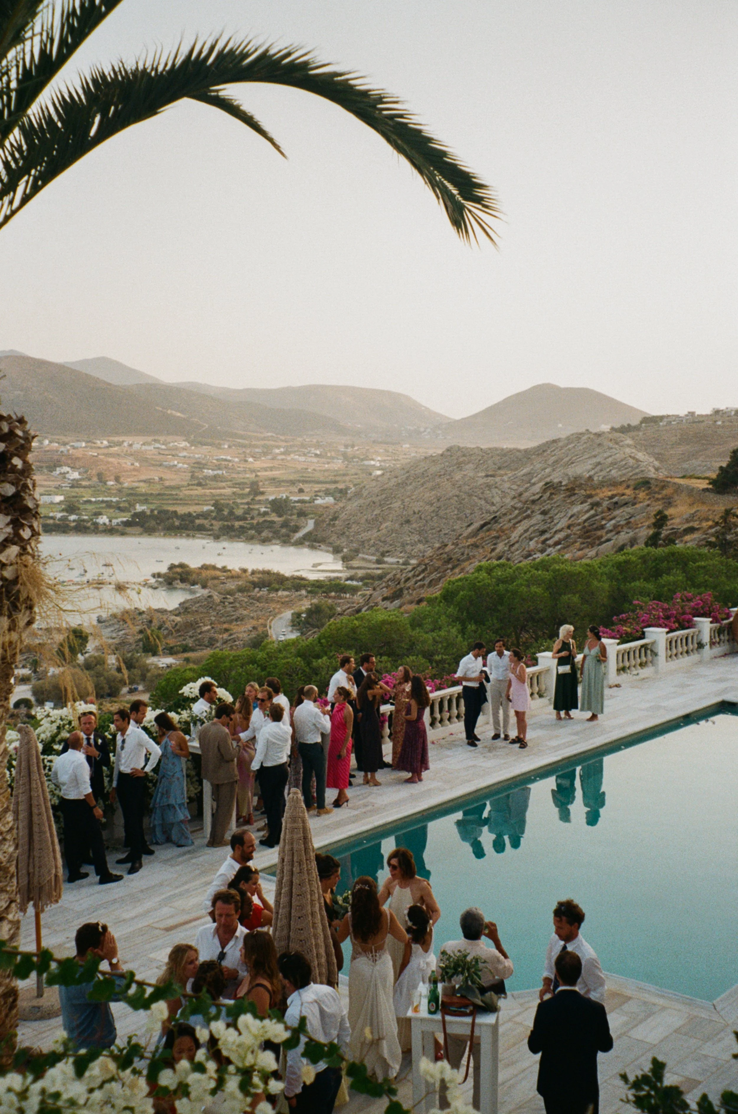 People gathered outdoors near a swimming pool, with hills and mountains in the background, during a social event or celebration.