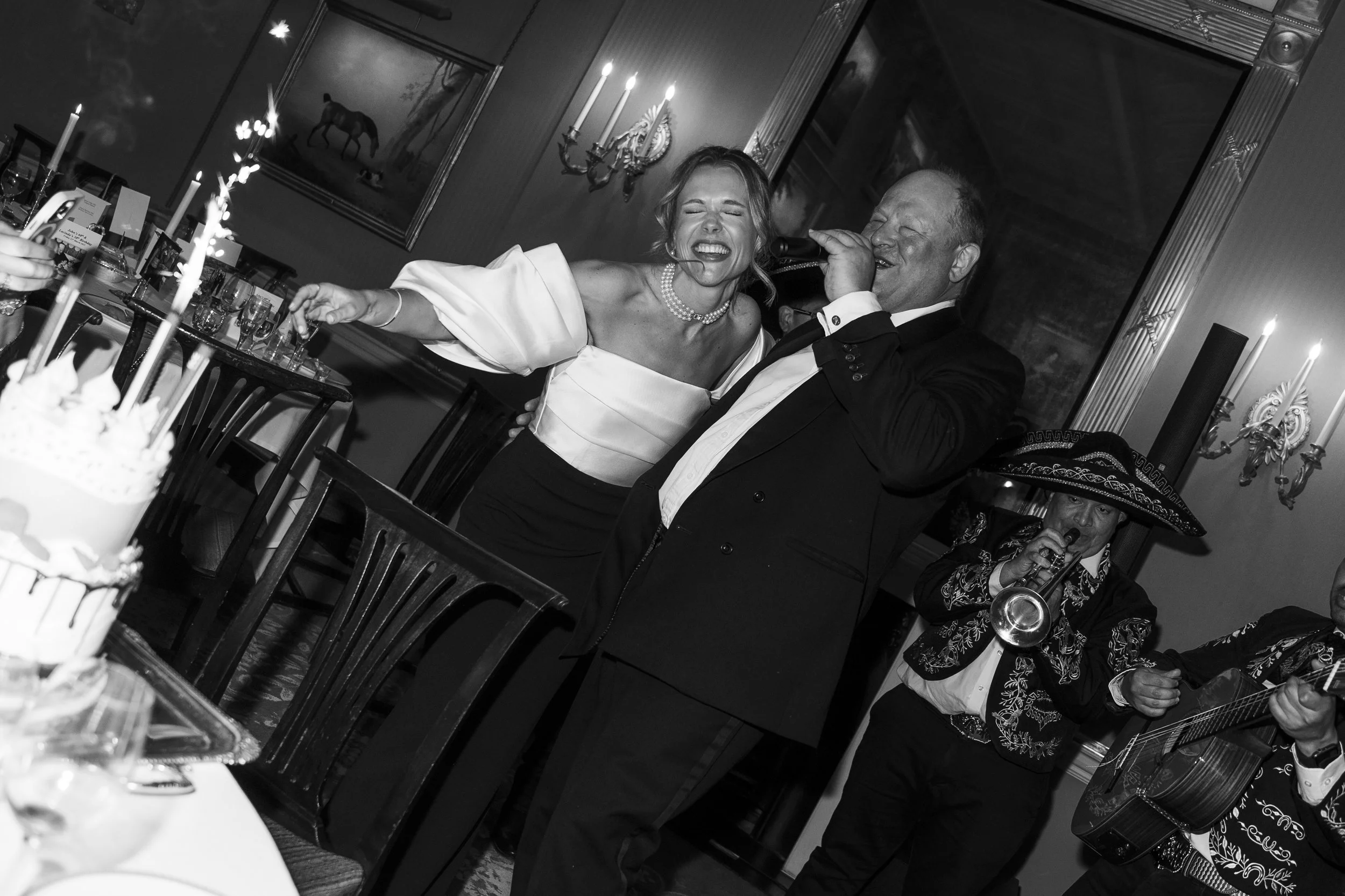 A woman in an off-shoulder dress and pearl necklace laughing and singing into a microphone, accompanied by an older man in a tuxedo. They are at a celebration with candles and drinks on the table, and other guests and a musician in a mariachi costume