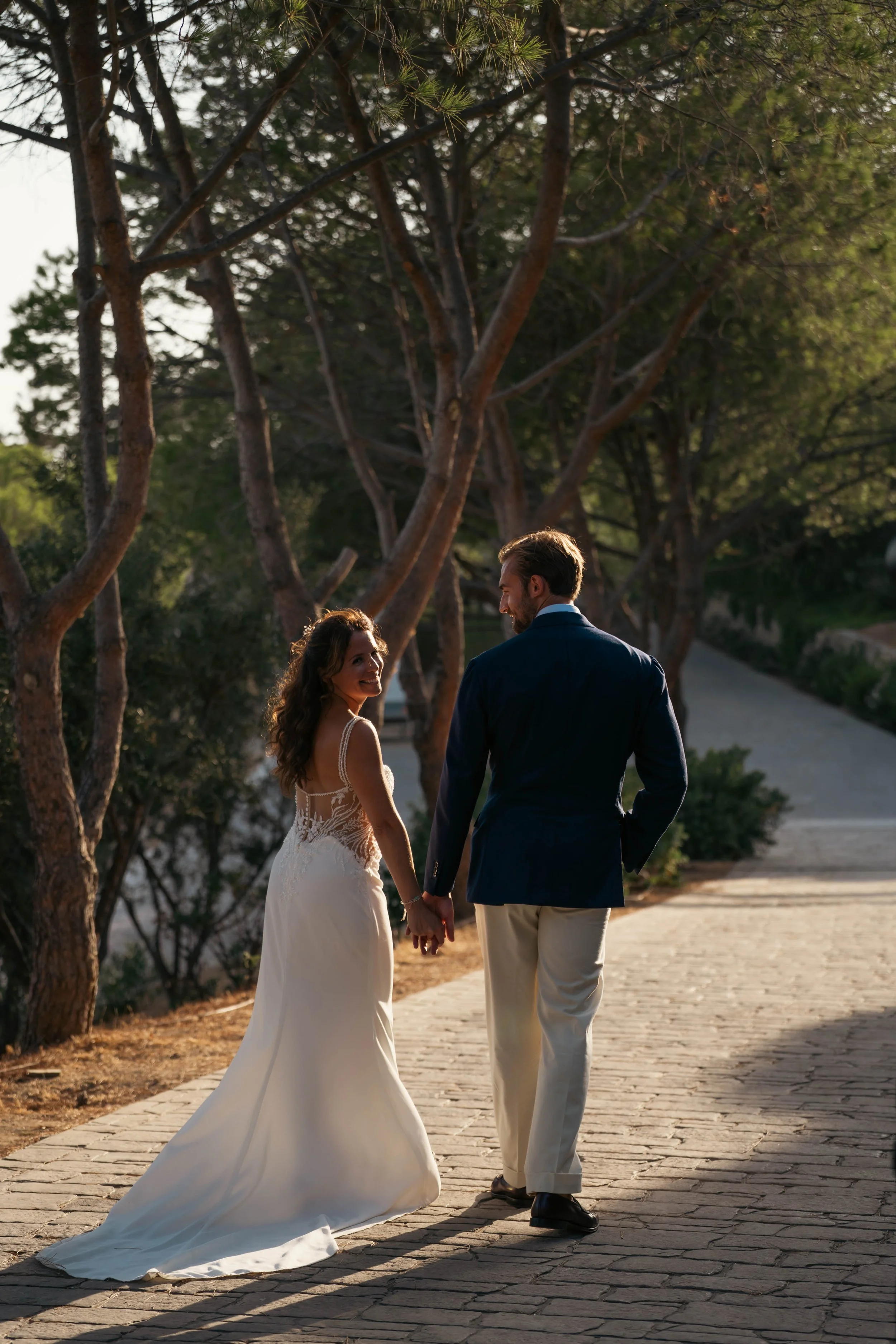 A bride and groom walking hand in hand on a paved pathway surrounded by trees during sunset.