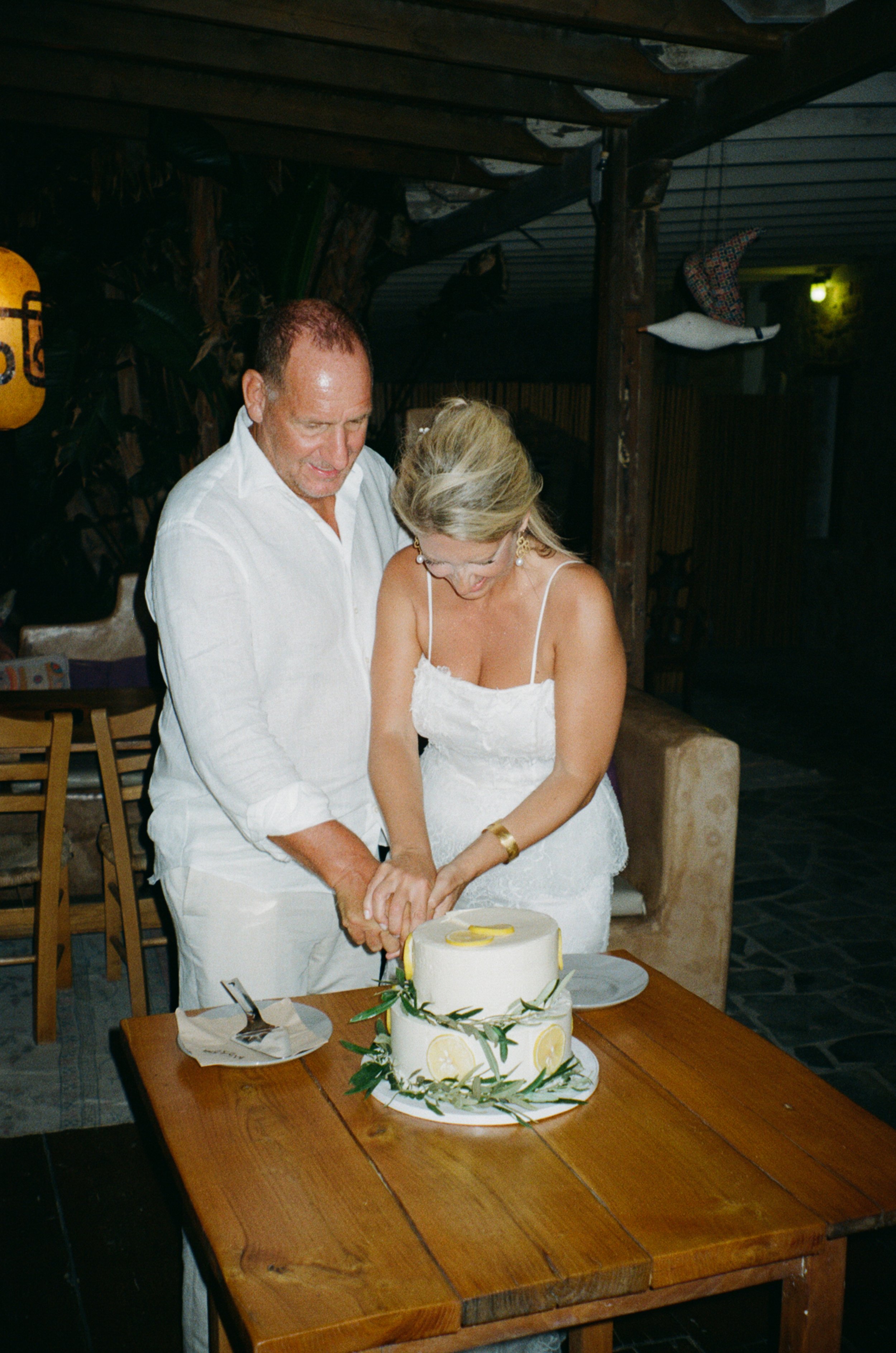 A man and woman, dressed in white, cut a wedding cake together at a celebration. The cake is decorated with lemon slices and greenery, placed on a wooden table with utensils and napkins. The setting appears to be a rustic indoor venue.