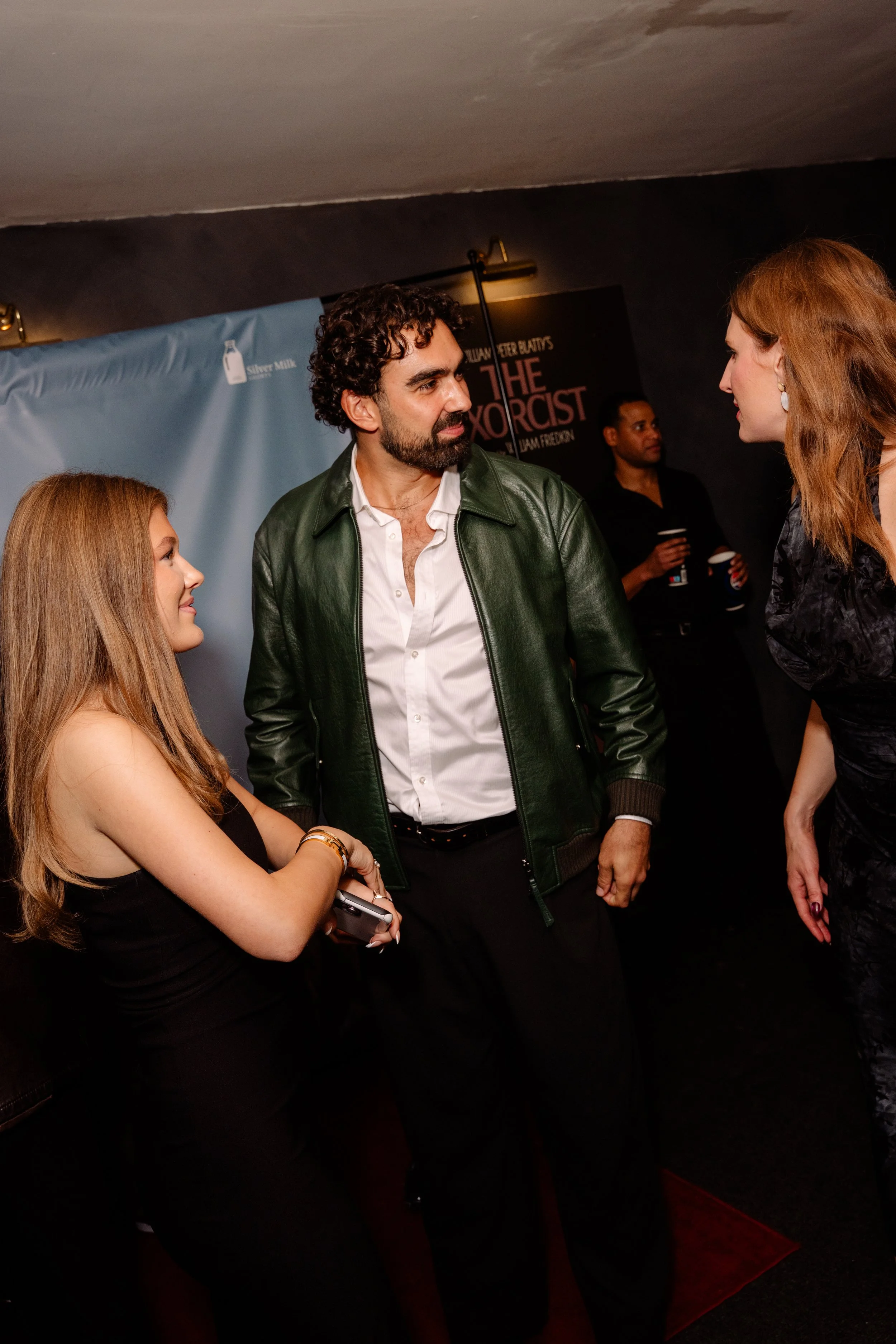 Four people engaged in conversation at a movie premiere or event, with a promotional poster for the film 'The Exorcist' visible in the background.