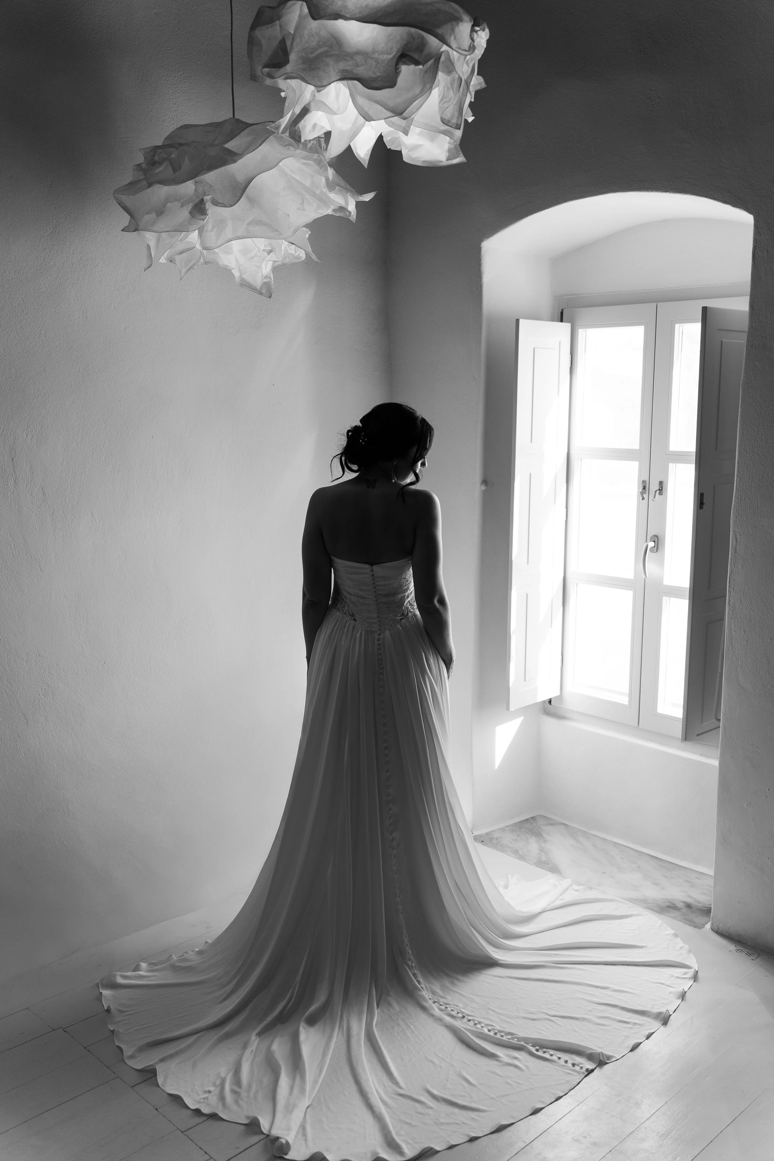 A woman in a wedding dress standing by an open window with sunlight streaming in, and crumpled paper pendant lights hanging from the ceiling, in a minimalist room.