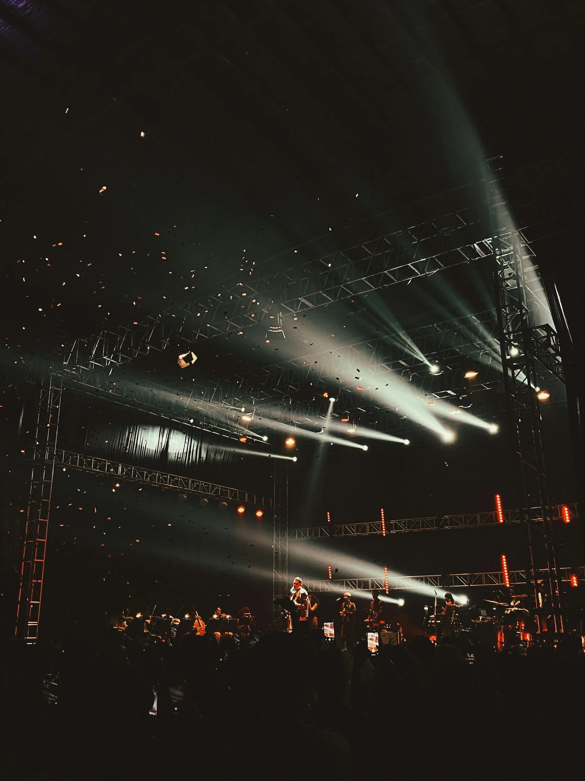 A concert stage with performers, illuminated by bright white spotlights, with structural metal scaffolding and a dark background.