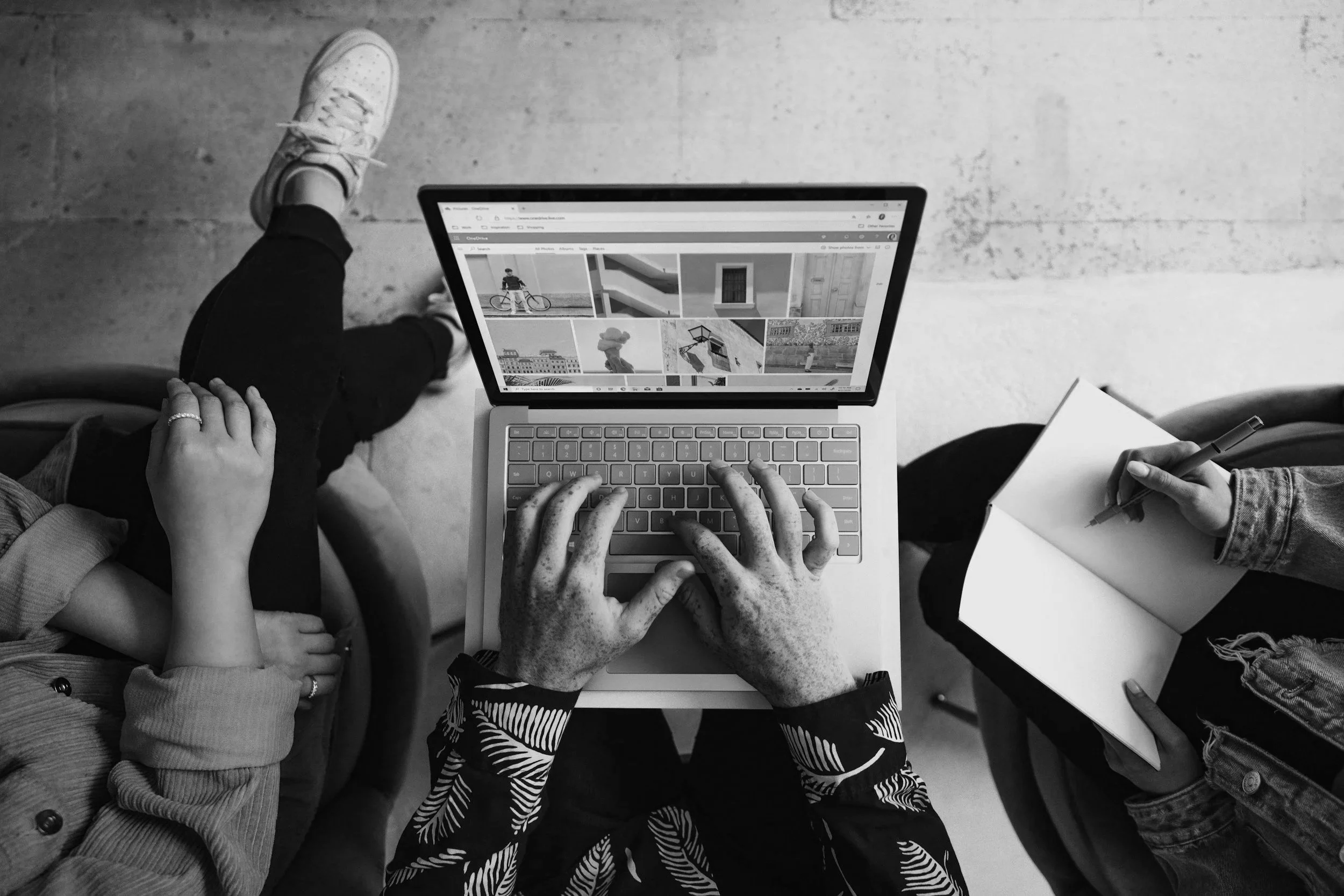 View from above of several people sitting around a laptop, one person is using the keyboard, another has a notebook and pen, and a third has their hand on the laptop. The laptop screen shows a grid of images. The scene is in black and white.