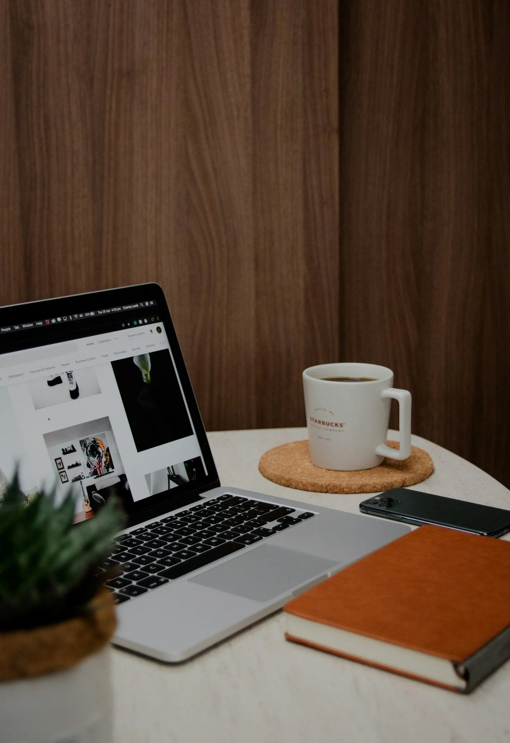 A workspace with a laptop showing a website, a coffee mug, a closed notebook, and a smartphone on a desk. In the foreground, part of a small green plant is visible, and the background features a wooden wall.