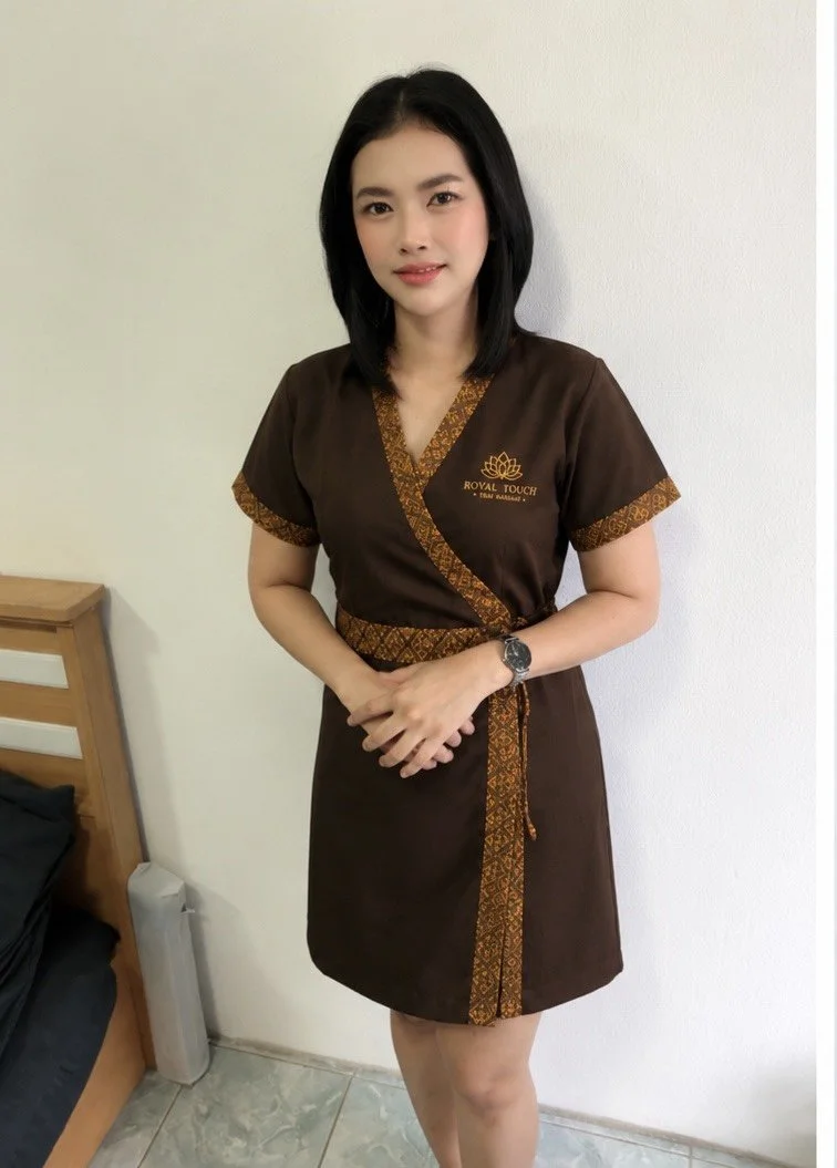 A young woman wearing a brown uniform with gold patterned accents, standing outdoors with lush green plants in the background, smiling and holding her hands in a prayer gesture.