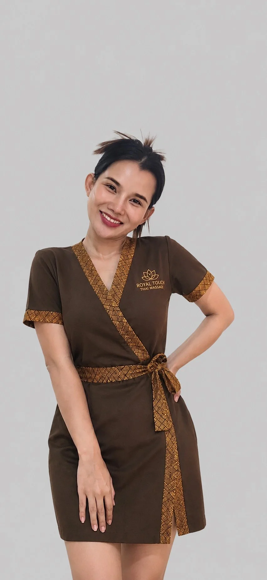 A woman wearing a brown uniform with gold accents and the logo 'Royal Touch Thai Massage' standing against a plain white background, smiling and posing with one hand on her hip.
