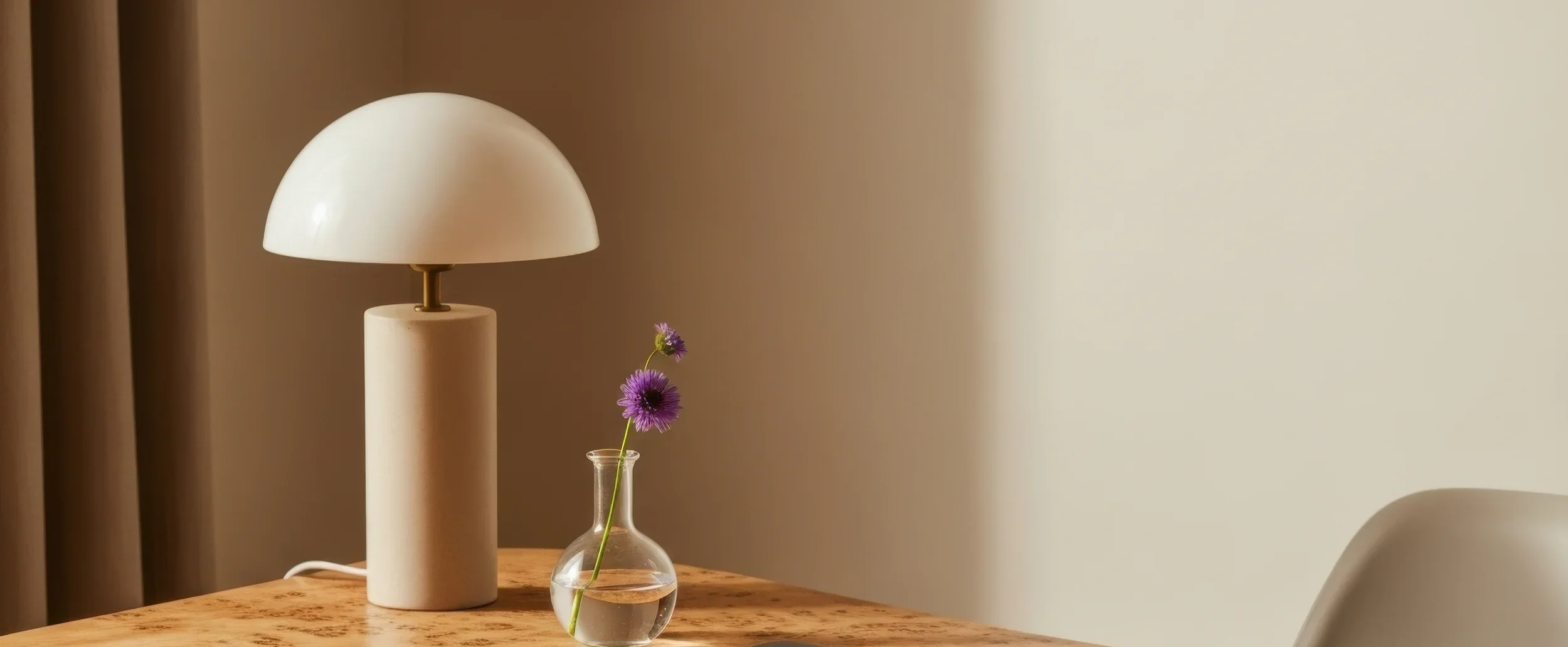 A table lamp with a white, semi-spherical shade and a beige cylindrical base. Next to the lamp, there is a small, clear glass vase with two purple flowers, one taller than the other. The scene is set in a room with a neutral wall and beige curtains in the background.