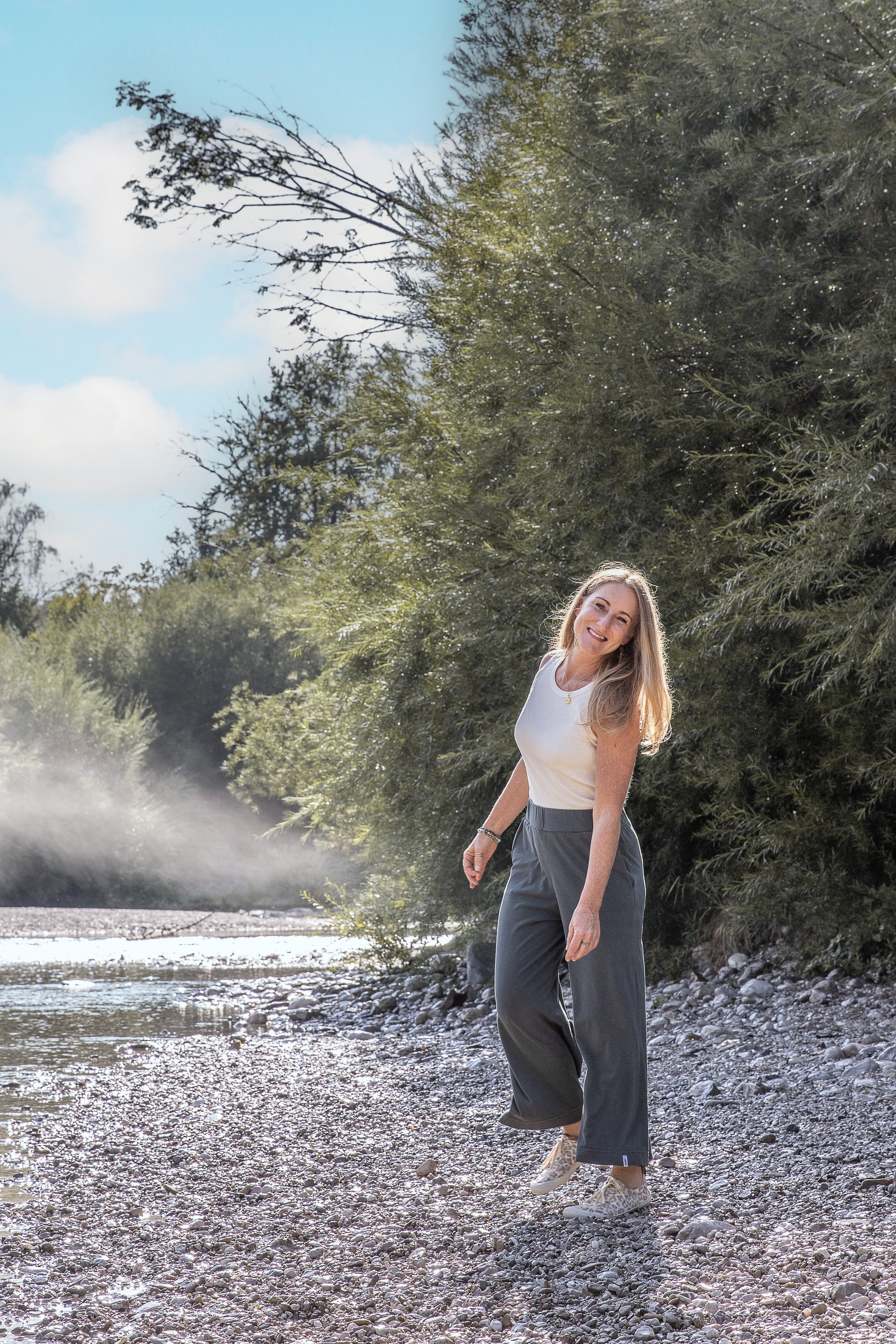 Eine junge Frau mit langen blonden Haaren, in grauen weiten Hosen, weißen Sneakers und einem weißen T-Shirt, steht am Flussufer in den Bergen, umgeben von grünen Büschen und Bäumen, bei Sonnenlicht, mit einem freundlichen Lächeln.