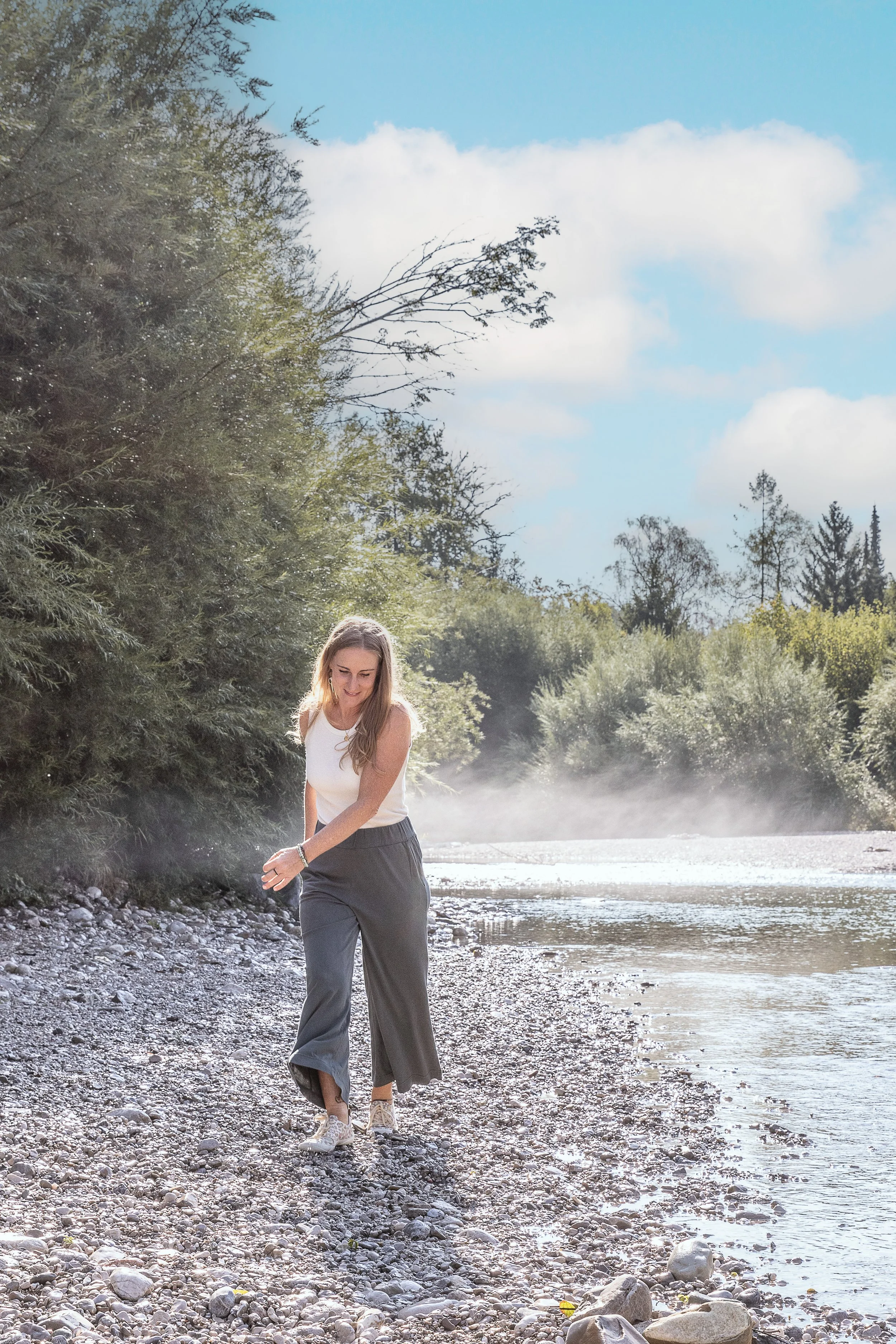 Eine junge Frau läuft barfuß an einem Fluss entlang, umgeben von Bäumen und blauen Himmel mit Wolken.