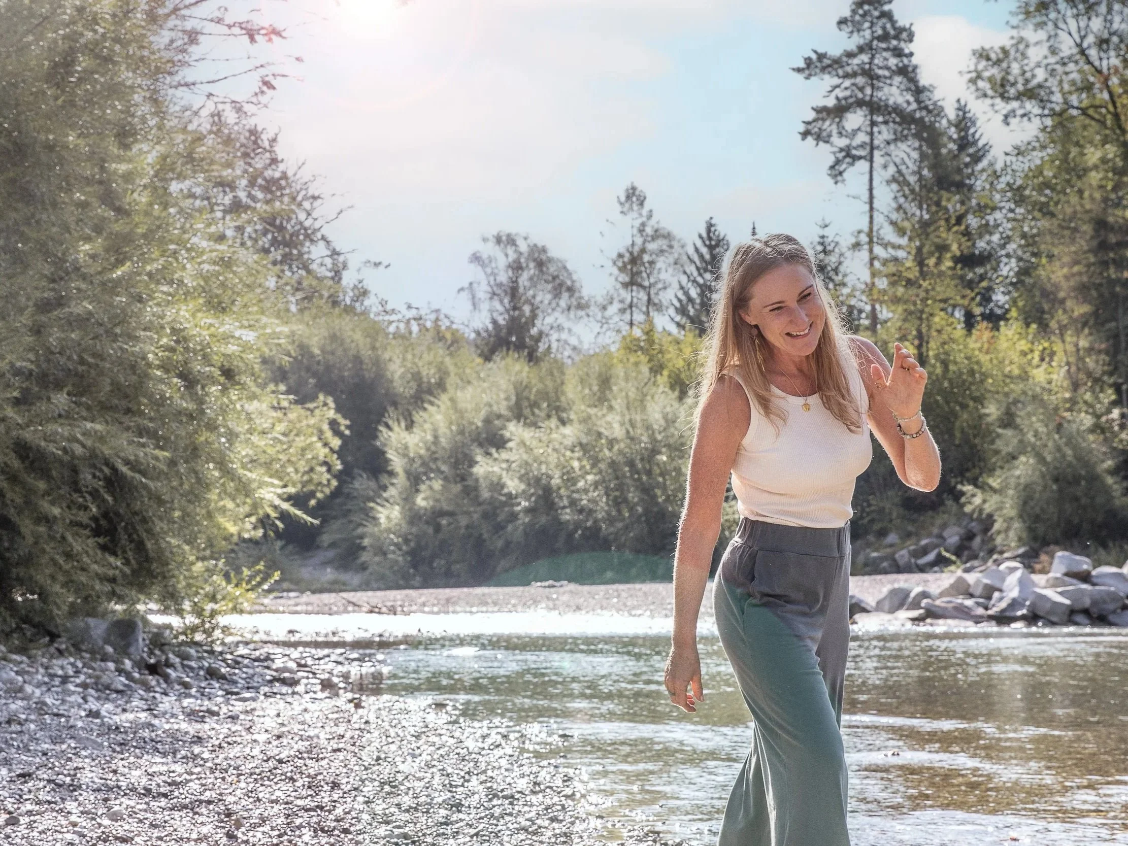 Eine lachende Frau in einer weißen Weste und grauen Hosen läuft an einem Fluss in einer bewaldeten Gegend.