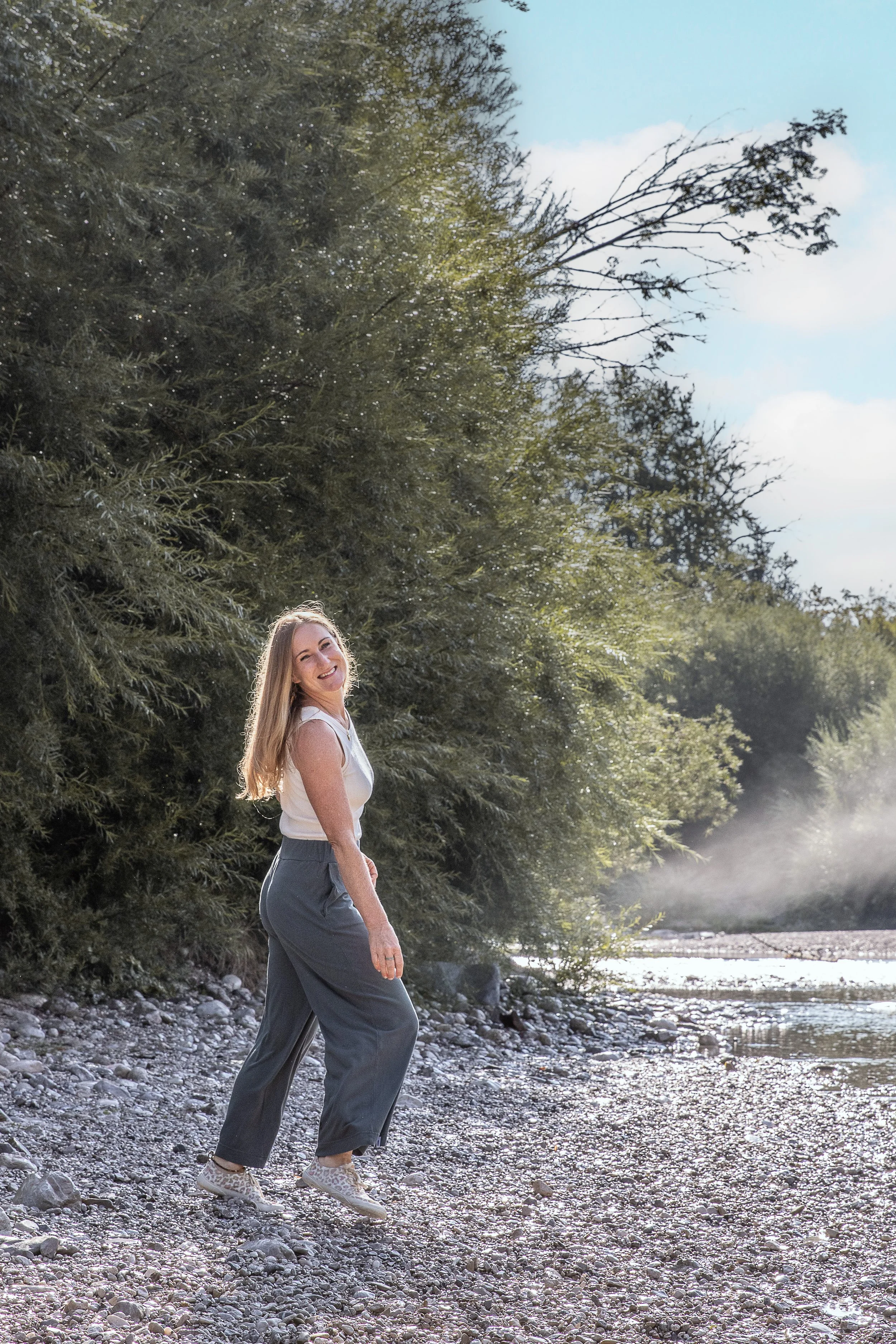 Junge Frau mit langem blondem Haar, lächelnd, stehend am Fluss, umgeben von grünen Büschen und Bäumen, bei Sonnenlicht, auf einem kiesbedeckten Flussufer.