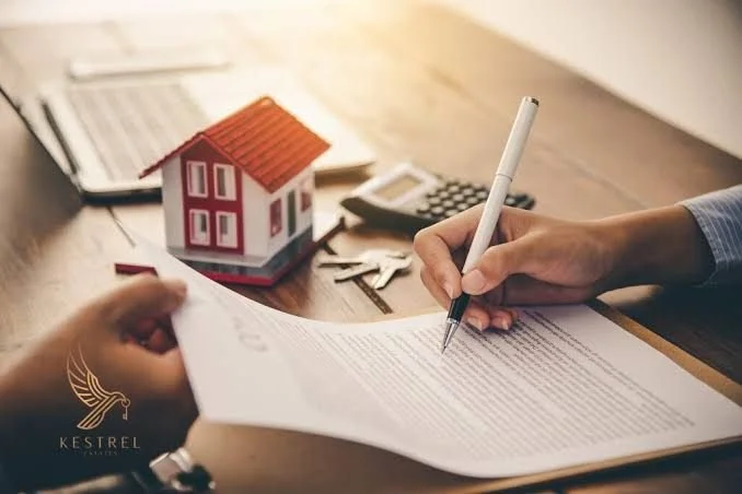 Person signing a document at a desk with a model house, calculator, and keys.