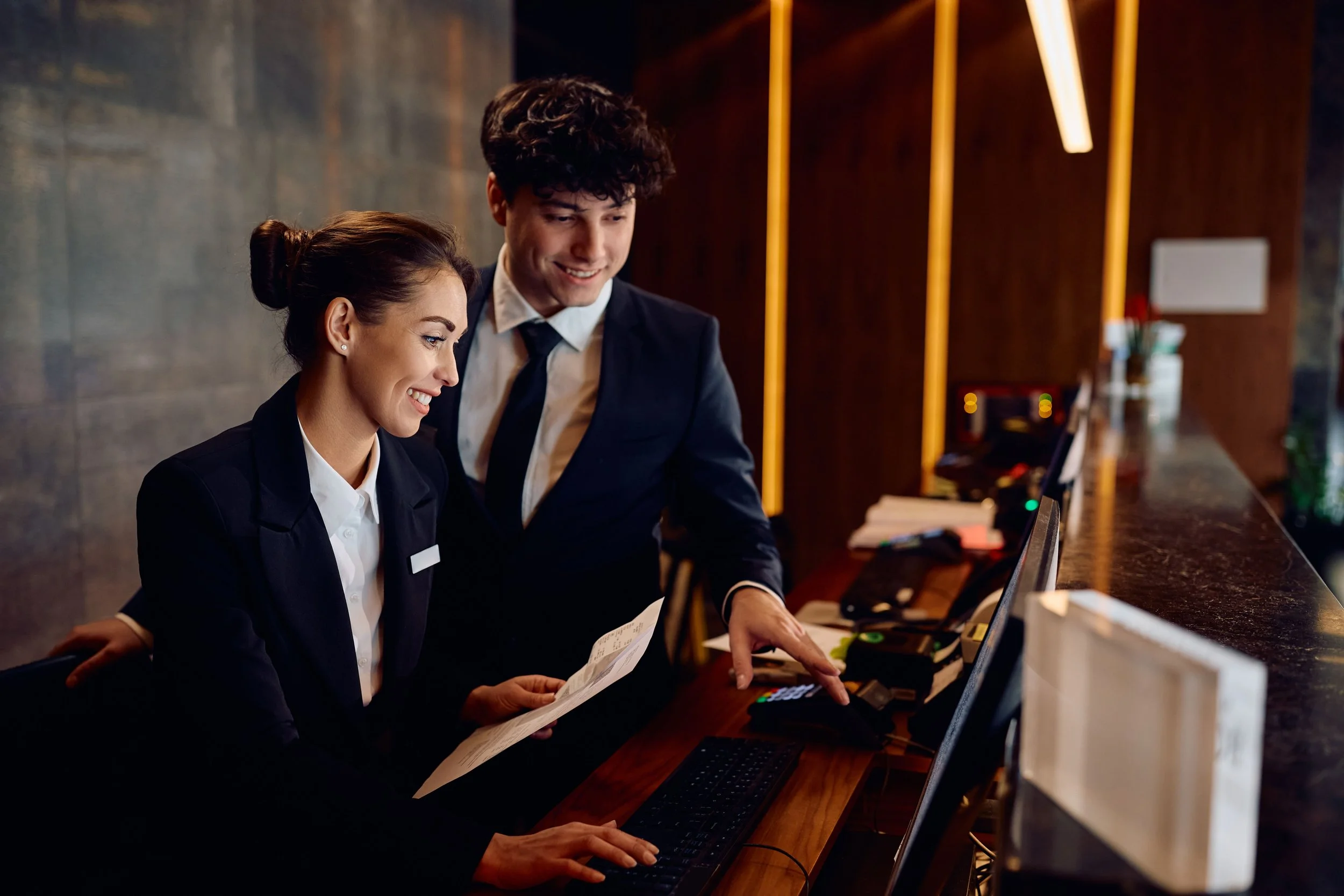 A woman and a man in formal business attire working together at a reception desk in a hotel or office setting, smiling and looking at documents or a check-in paper.
