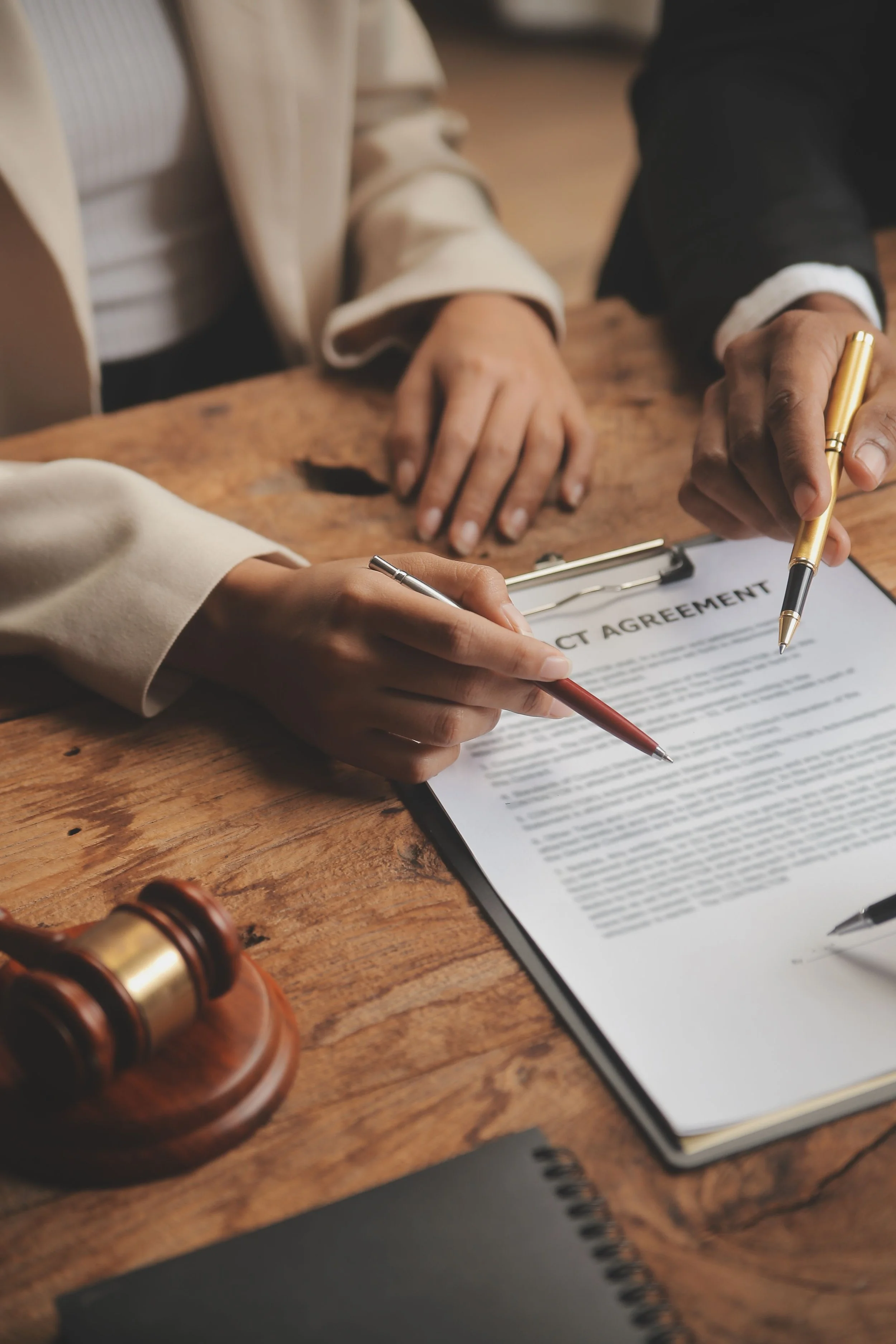 A close-up of a legal contract on a clipboard being signed by two individuals, one with a gold pen and the other holding a silver pen, on a wooden table with a judge's gavel in the foreground.