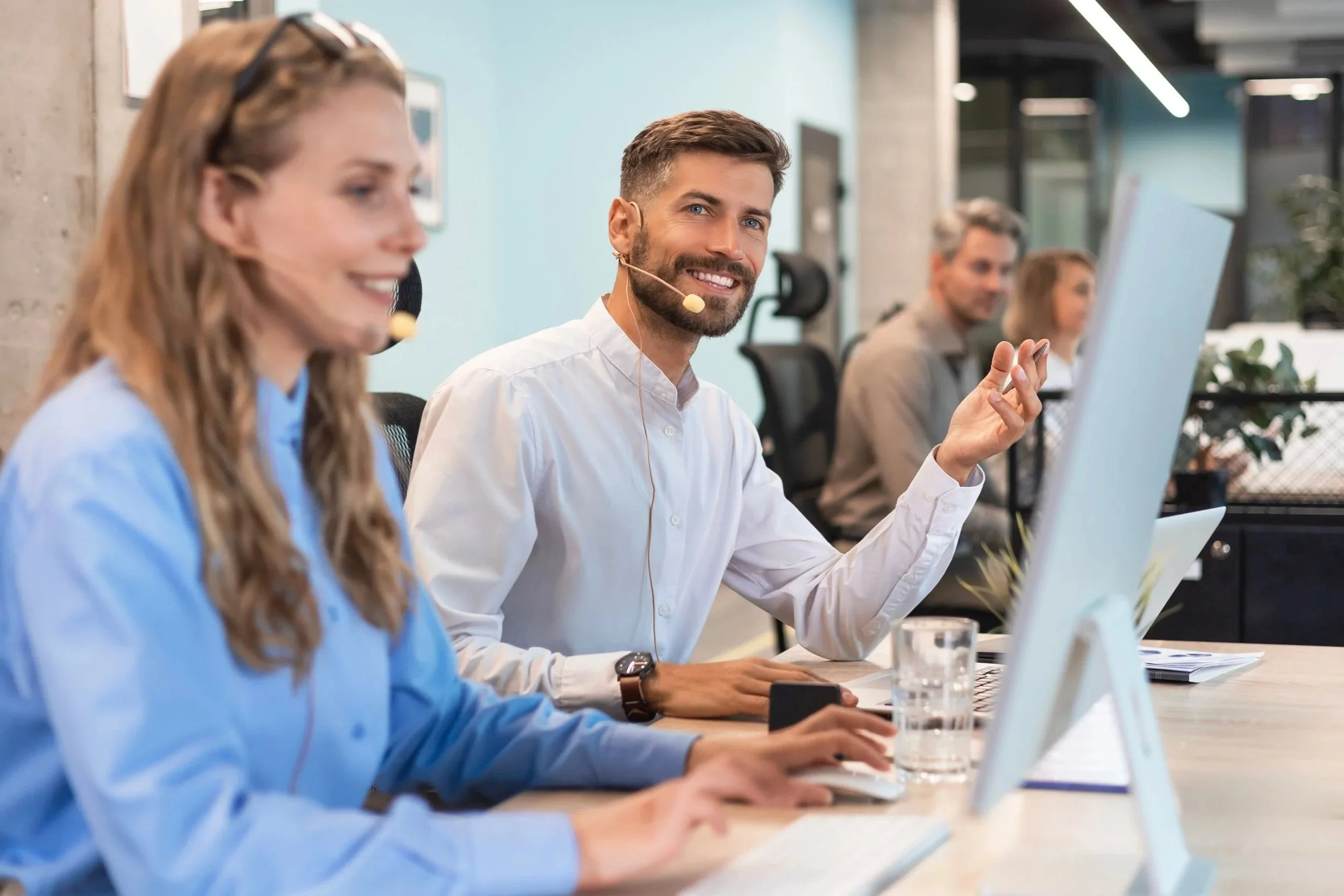 People working at a long table in a modern office, with a man smiling and speaking into a headset, and others working on computers.