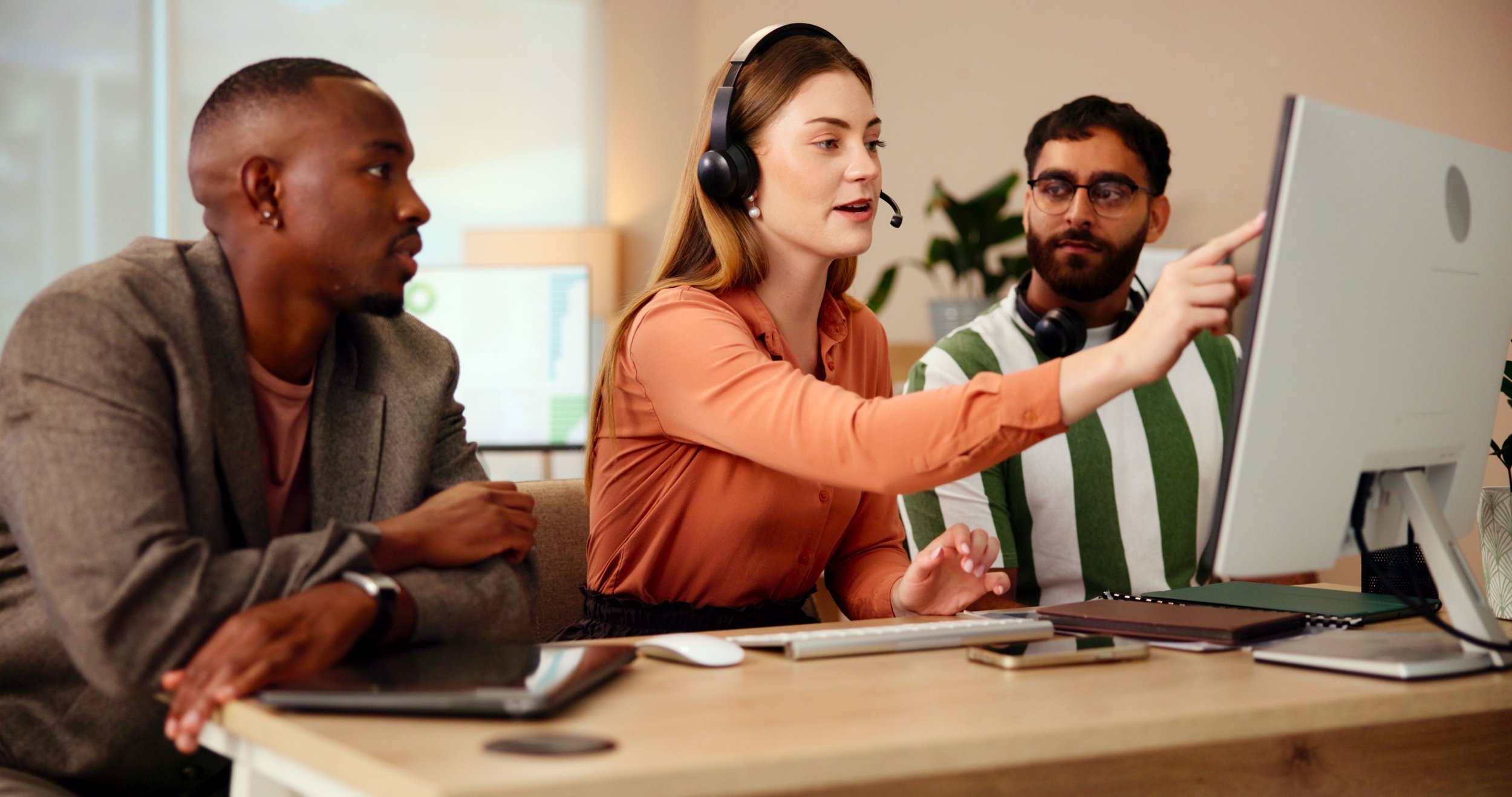 Three people working together at a desk in an office. The woman in the middle is wearing a headset and pointing at the computer monitor while speaking to her colleagues. The man on the right is looking at the monitor, and the man on the left is also focused on the conversation and the screen.