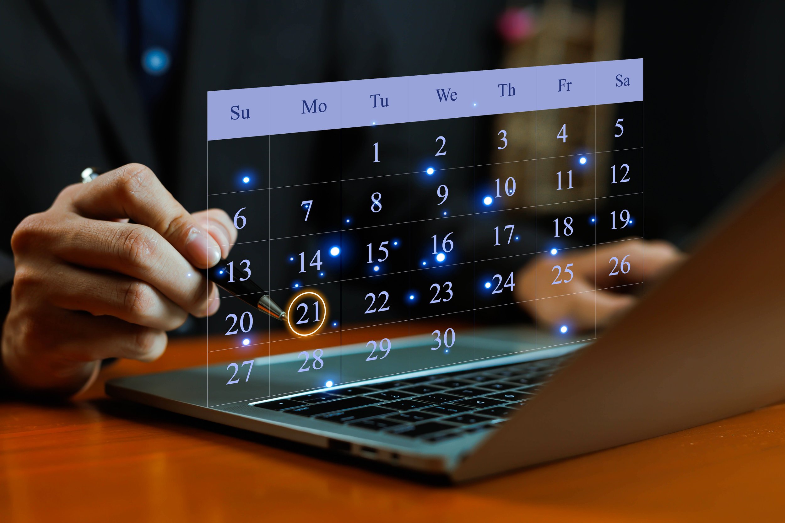 A person holding a pen over a digital transparent calendar displaying dates between the 1st and 30th, with some dates glowing. The calendar is projected above a laptop keyboard on a wooden desk.