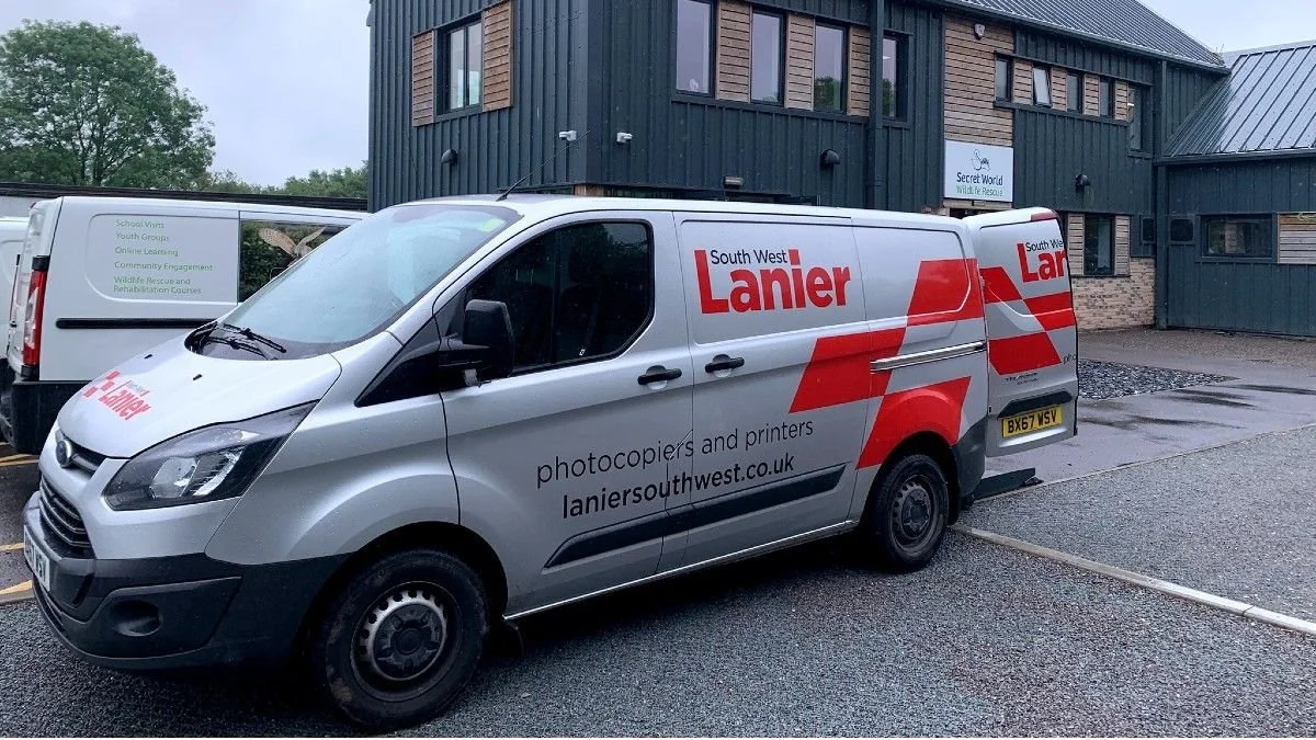 A silver van with red and black branding for South West Lanier, parked outside a dark green building with wooden accents and a sign for Secret World Wildlife Rescue.