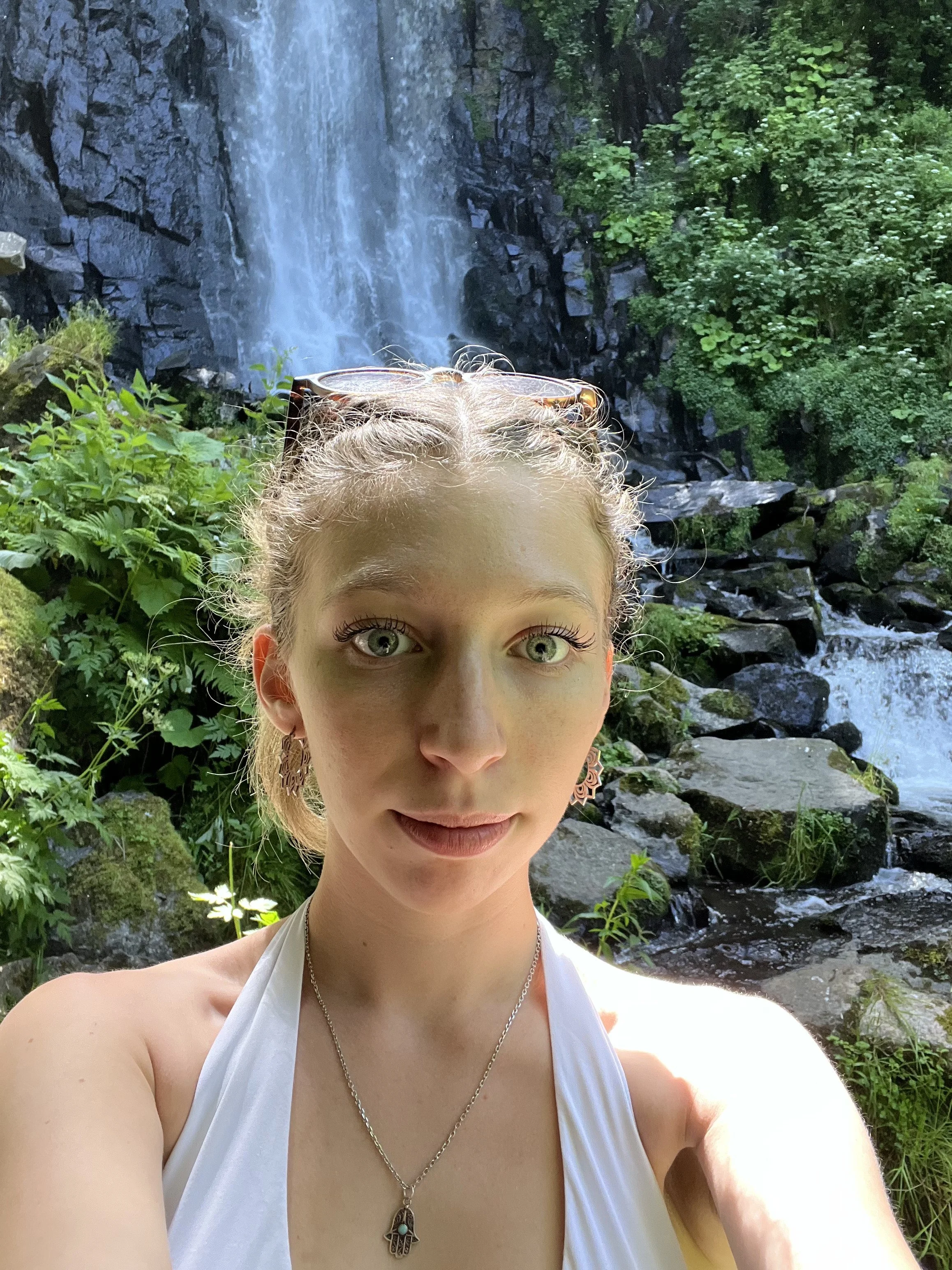 Une femme avec des cheveux blonds, portant des boucles d'oreilles, un collier avec un pendentif en forme de main de Fatma, et une robe blanche, se tient devant une cascade dans une forêt dense.