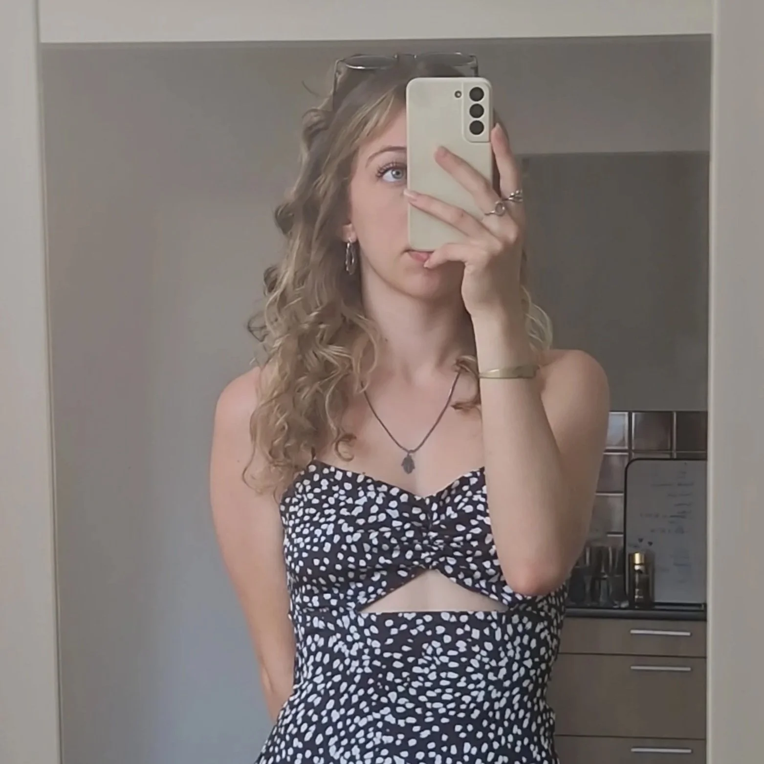 Une jeune femme avec des cheveux bouclés, portant un haut sans manches à motifs noirs et blancs, prenant un selfie dans un miroir, avec des lunettes sur la tête et des accessoires en argent. Elle se trouve dans une cuisine moderne.