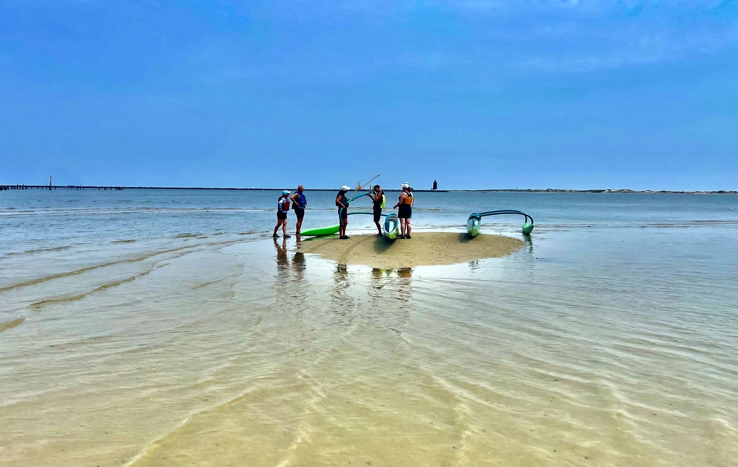 A group of six people preparing to go paddleboarding on a beach with clear water and a pier in the background.