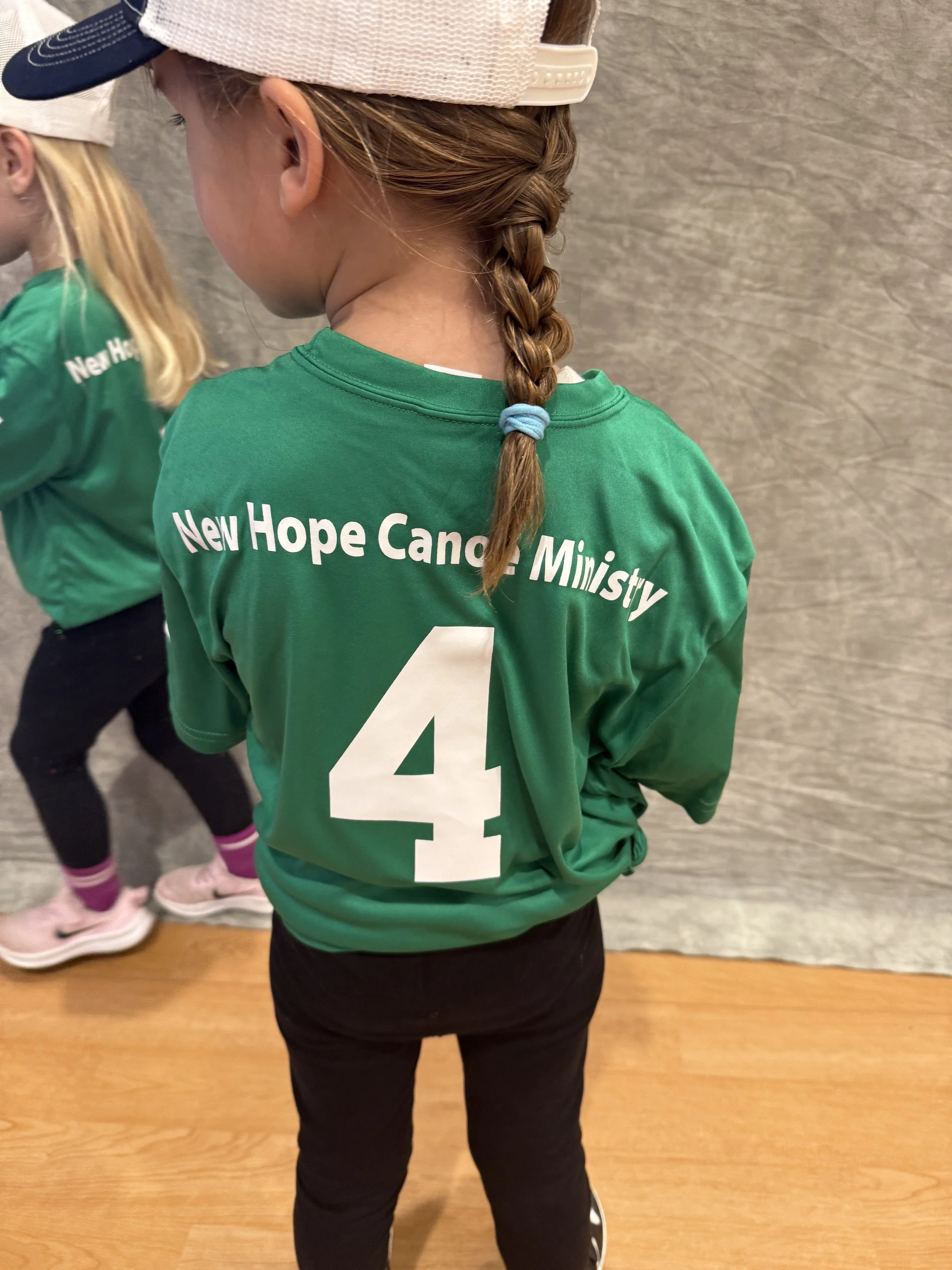 Child wearing a green sports jersey with the words "New Hope Cancer Ministry" and the number 4, standing indoors with a wooden floor and a textured wall in the background.