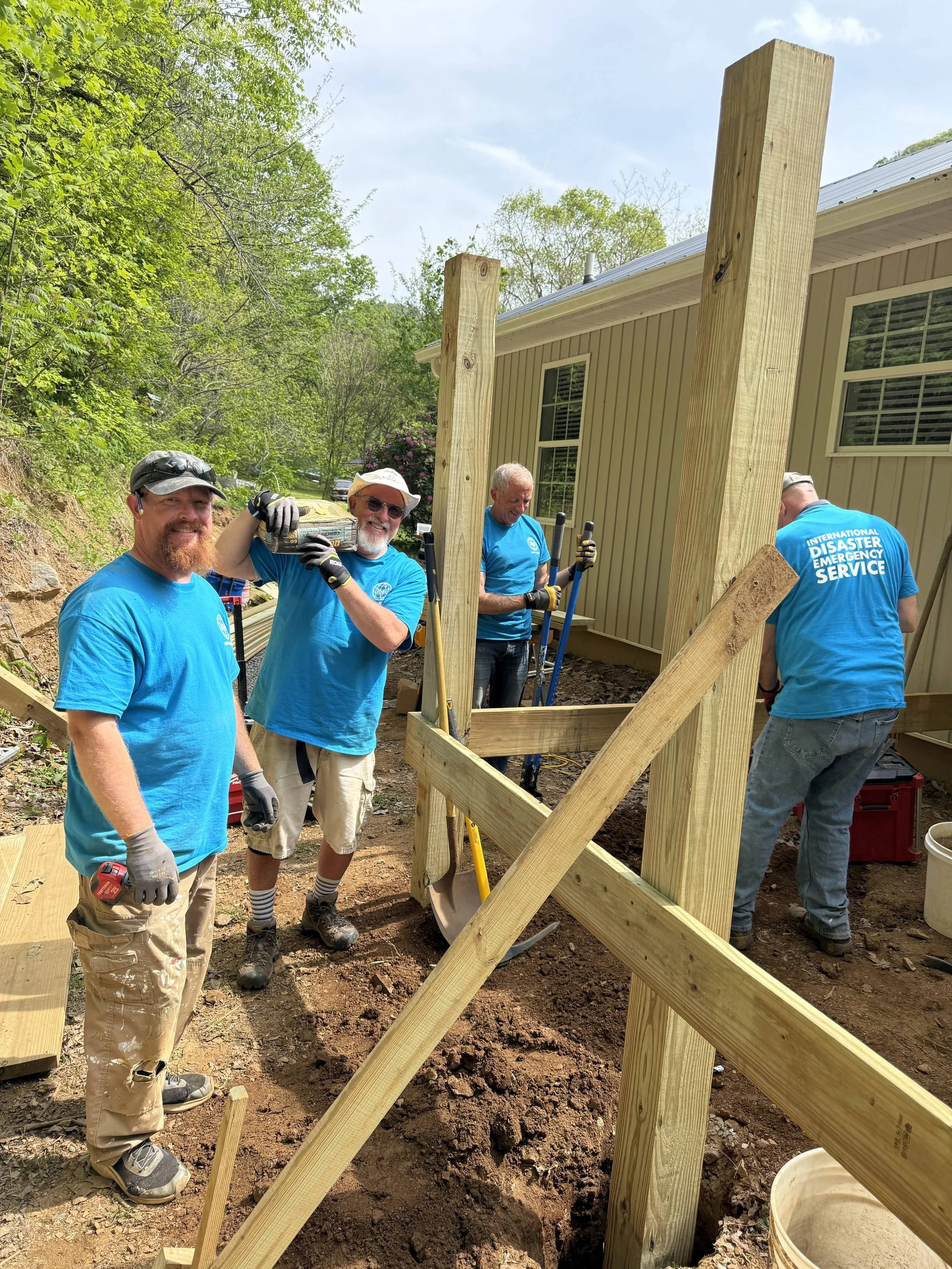 Four men working on a construction project outdoors, building a wooden structure near a house with beige siding. One man is smiling at the camera, holding a power drill, while the others are focused on the construction work. They are wearing blue shirts and gloves, surrounded by tools and wood. Trees and a blue sky are in the background.